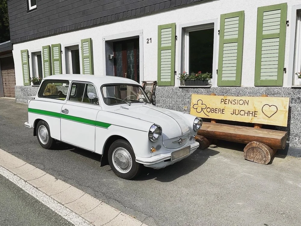 Ein weißer Oldtimer mit grüner Seitenlinie steht vor einem Haus mit grünen Fensterläden. Es gibt einen Holzbank mit einem Schild, das 'PENSION OBERE JUCHHE' und eine Herzzeichnung zeigt.