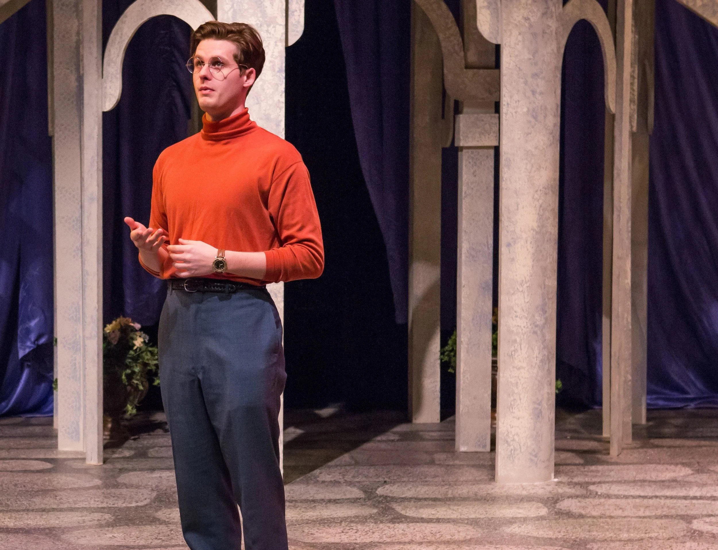 Male actor in orange turtleneck on stage with columns and dark curtains