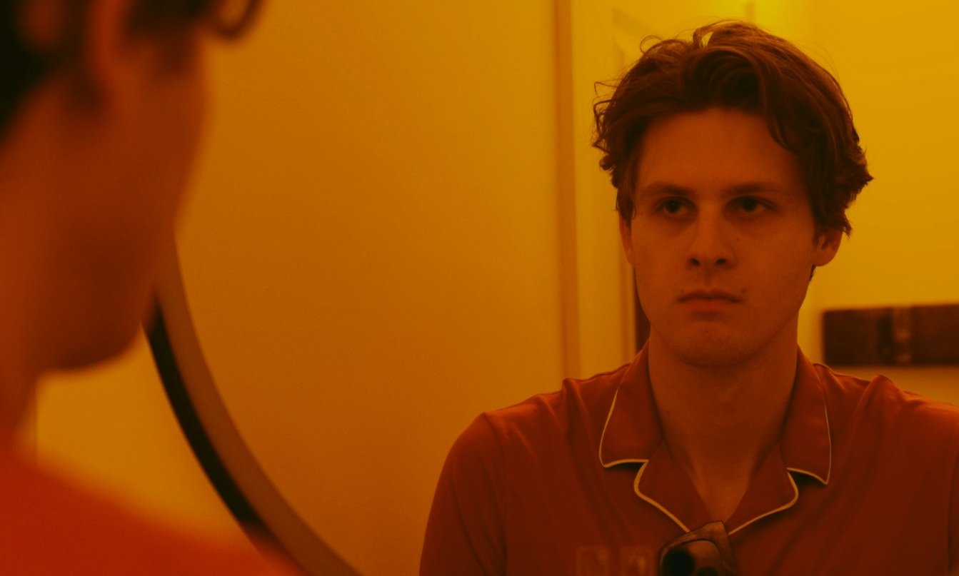 A man looking at himself in a mirror, wearing a red shirt, with a serious expression, in a warmly lit room.