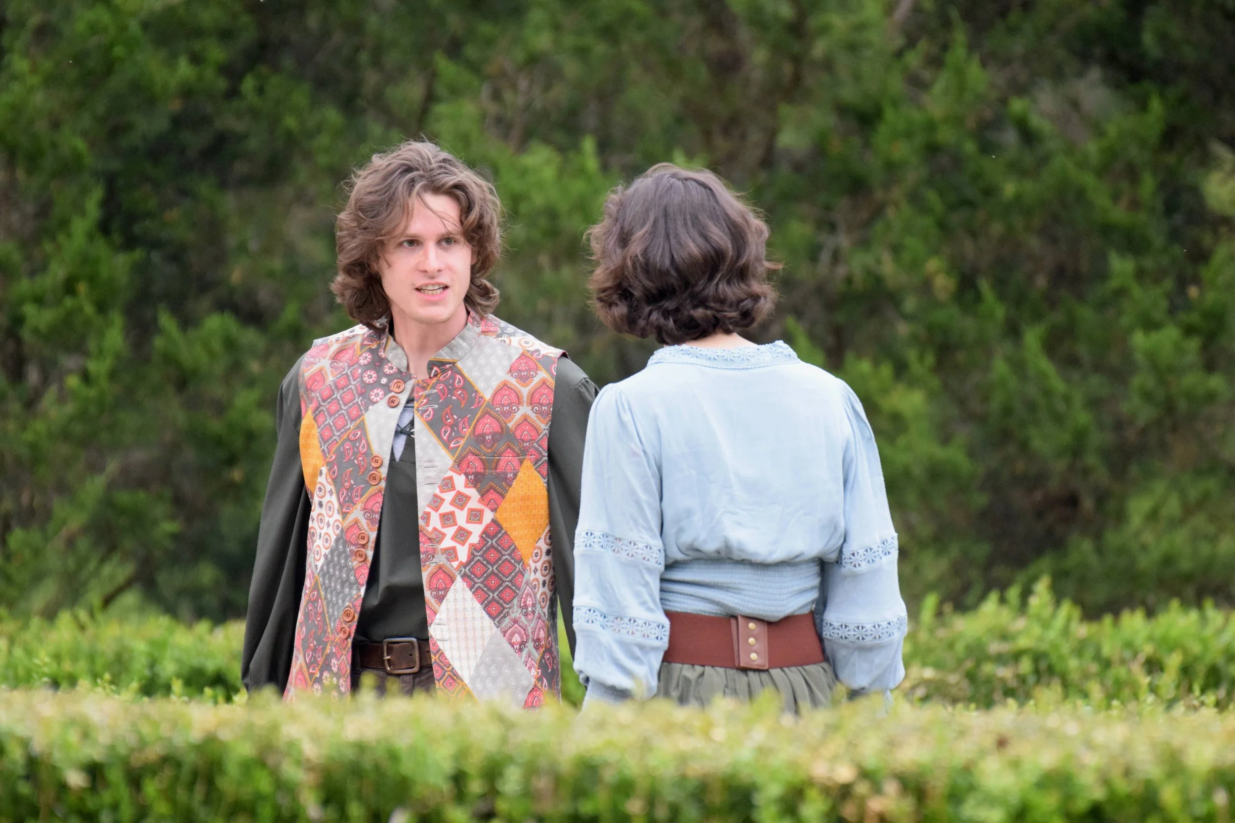 Two people dressed in medieval-style clothing stand outside in a garden area with one facing the camera and the other turned away.
