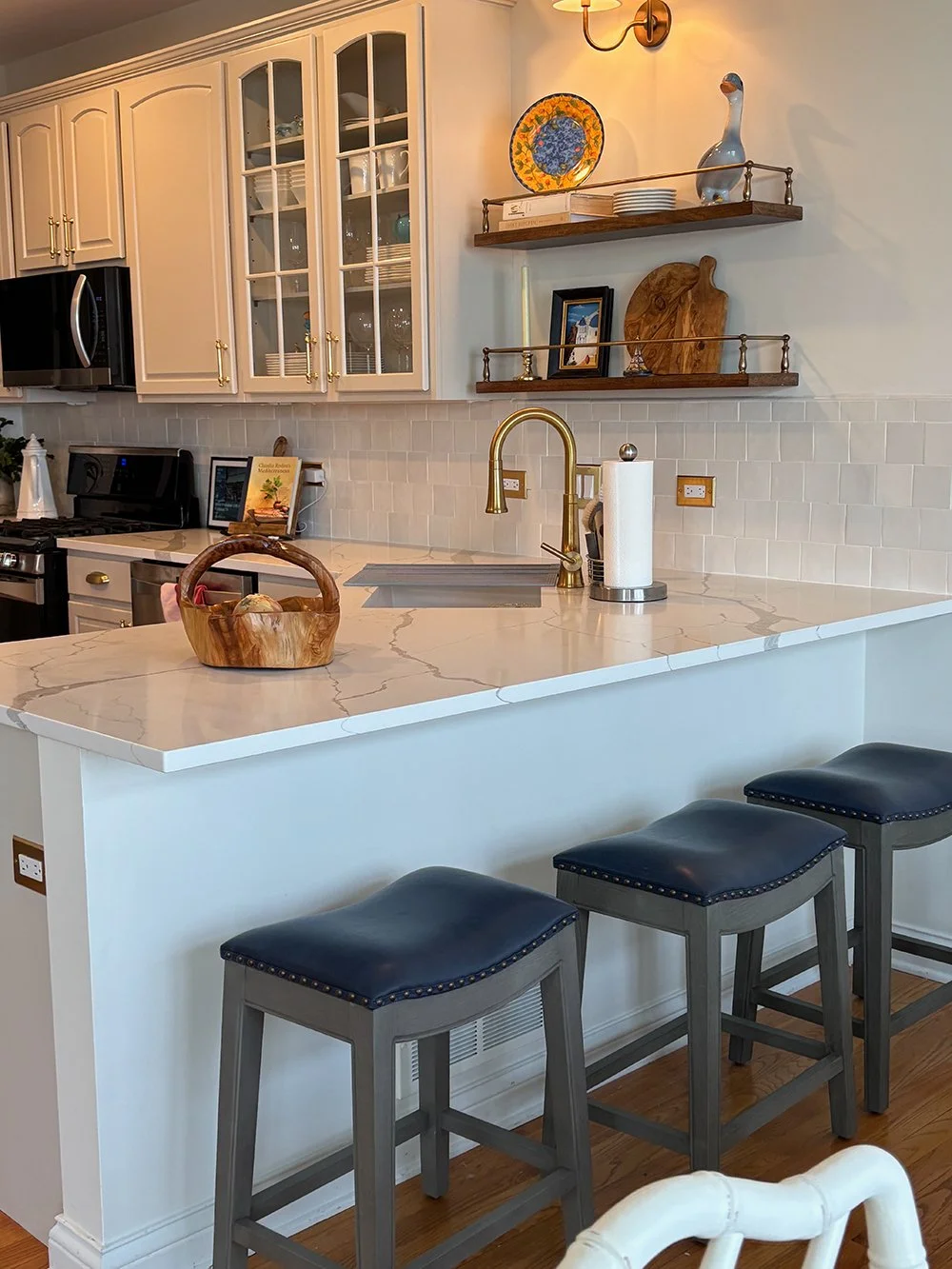 A kitchen with white cabinets, a marble countertop, a gold faucet, and three blue cushioned bar stools. Open shelves hold decorative items like a plate, a sculpture, and picture frames. There is also a microwave, dishwasher, and stove.