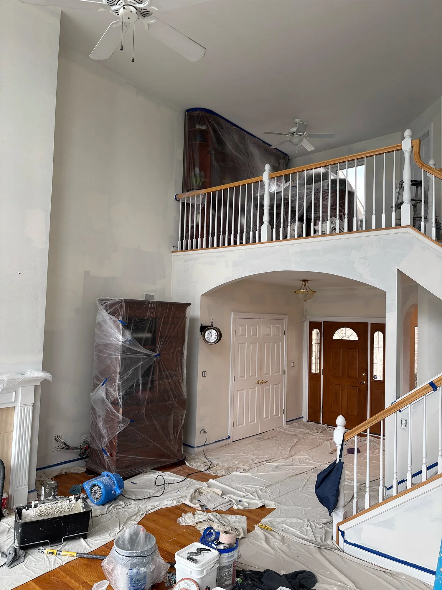 Living room in the process of painting or renovation, with plastic covering furniture, paint supplies on the floor, and construction tools scattered around.