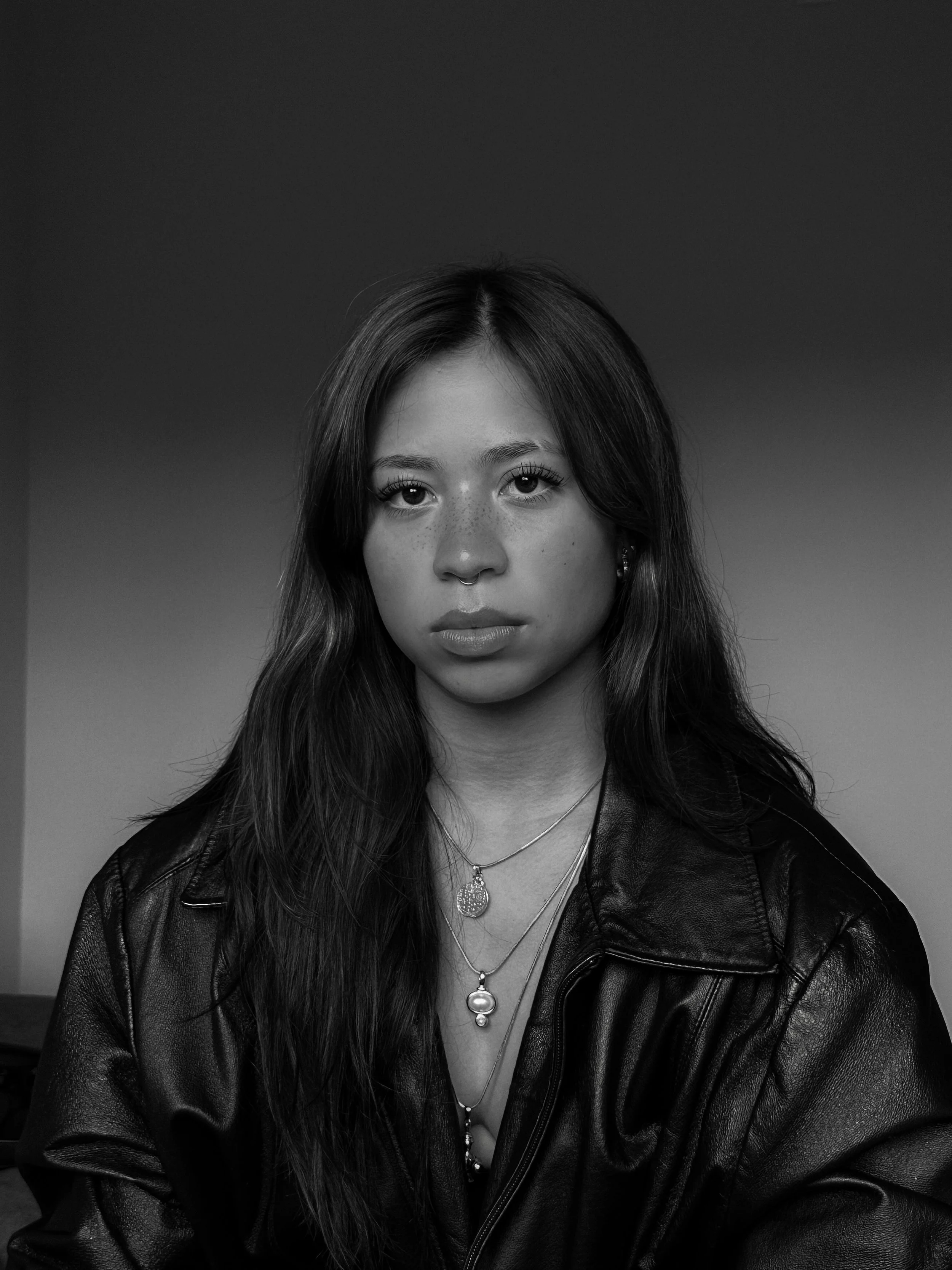 Black and white portrait of a young woman with long hair, wearing layered necklaces and a leather jacket, looking directly at the camera.