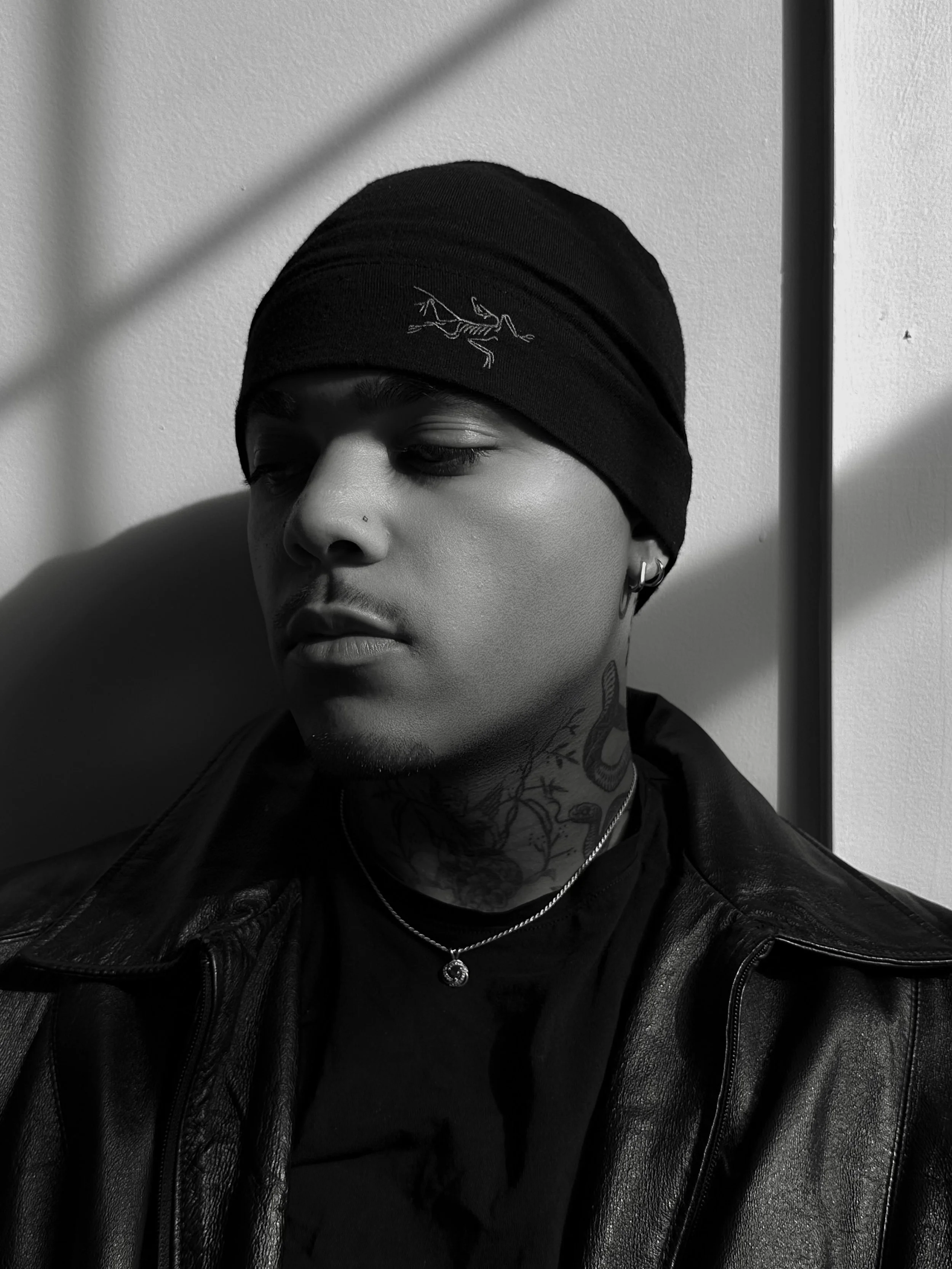 A young man with tattoos on his neck, wearing hoop earrings, a necklace, a black leather jacket, and a black beanie with a logo, leaning against a wall with shadows cast on his face.