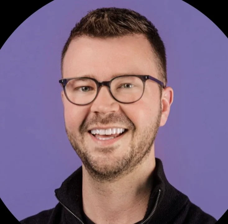 A smiling man with short brown hair, glasses, and a beard, wearing a black zip-up jacket against a purple background.