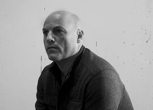 A black and white photograph of a person with shoulder-length hair looking to the side, with colorful lens flare effects.