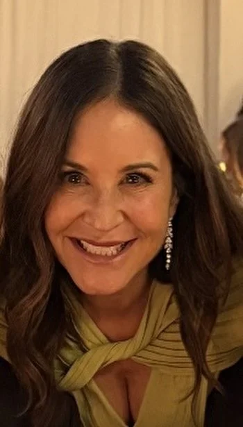 A woman smiling, with long brown hair, wearing a yellow top with a knot detail and large, dangling earrings.