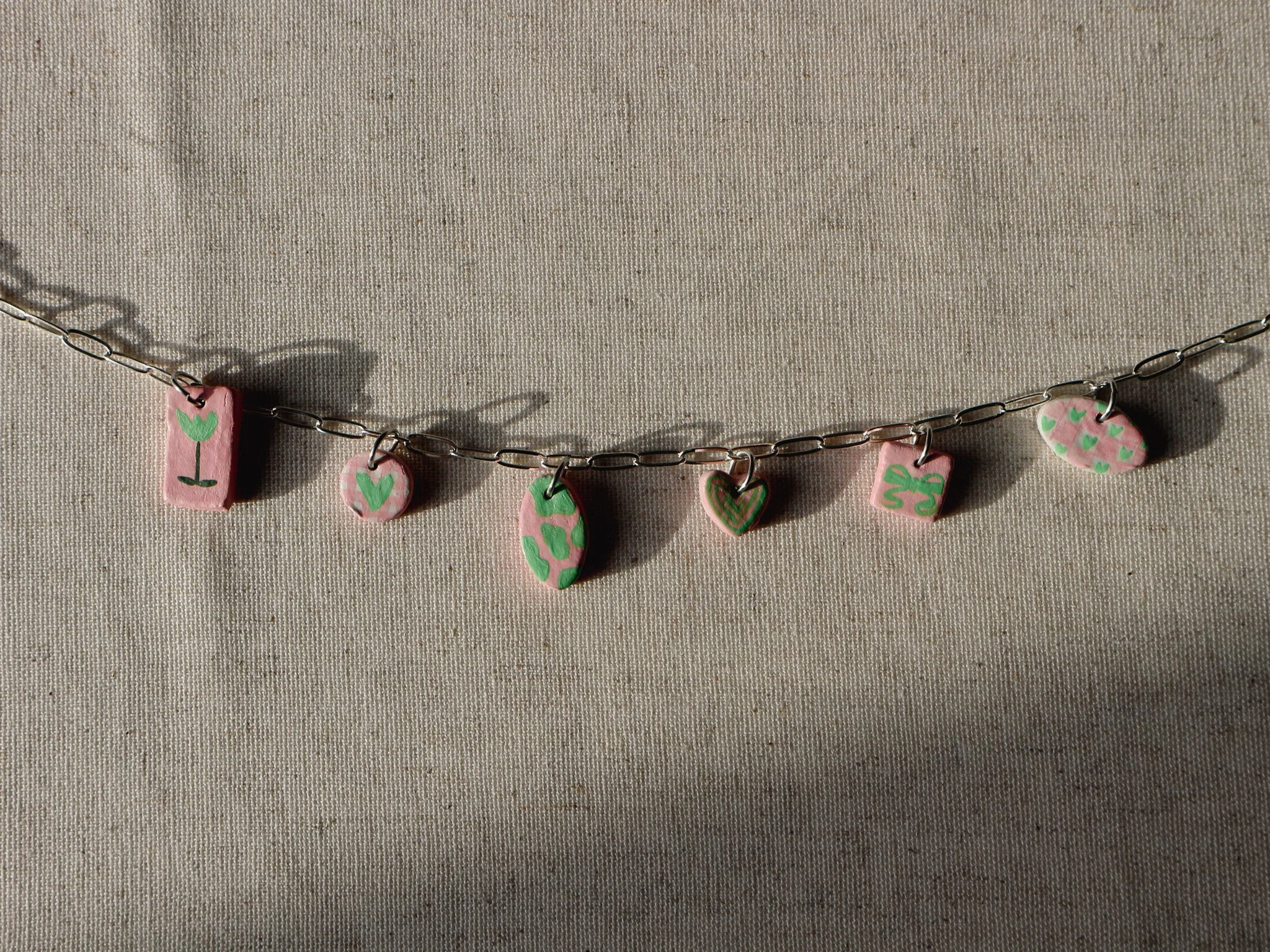 A necklace with pink and green charms, each featuring various green heart and flower designs on a thin metal chain, against a light beige fabric background.