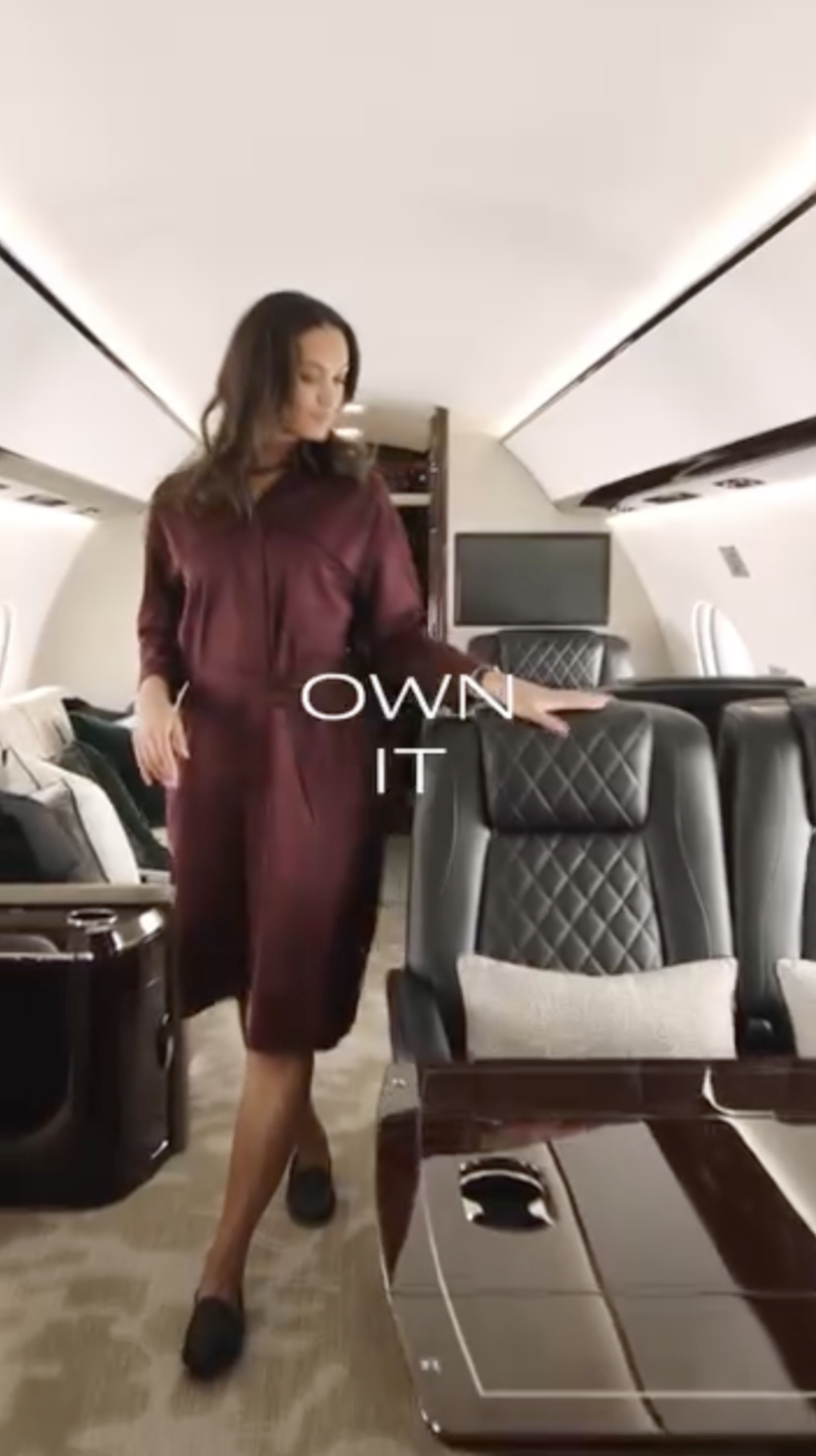 Woman walking through a Gulfstream aircraft cabin with her hand on the back of a seat and on-screen text that reads, "Own it."