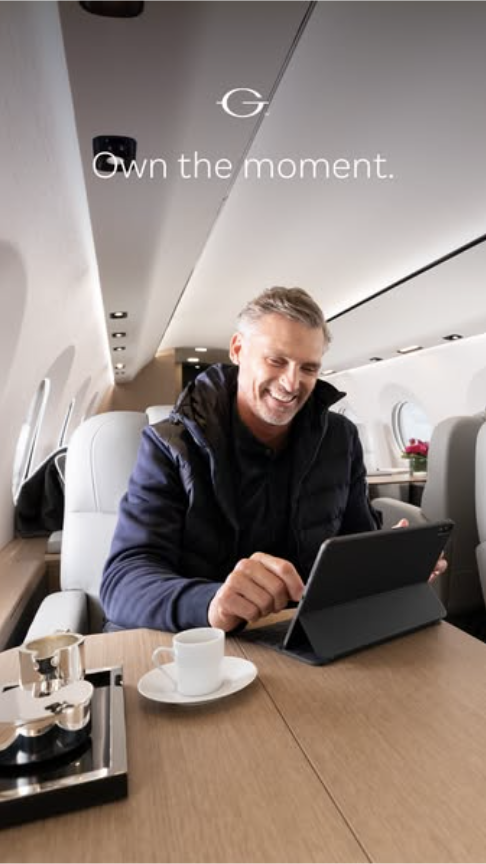 Smiling man sitting inside a Gulfstream airplane working on a computer and drinking coffee, with on-screen text that reads: "Own the moment."
