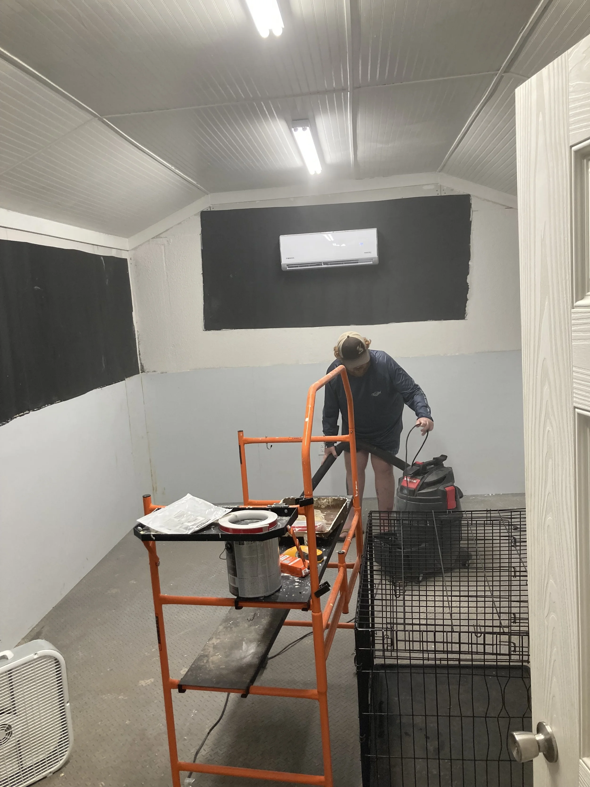 A person wearing a dark jacket and shorts is working on cleaning in a small room, with a vacuum cleaner. The room has a white ceiling with fluorescent lights, dark walls, and a black air conditioning unit. There is equipment and a wire basket in the room, with a fan on the side.