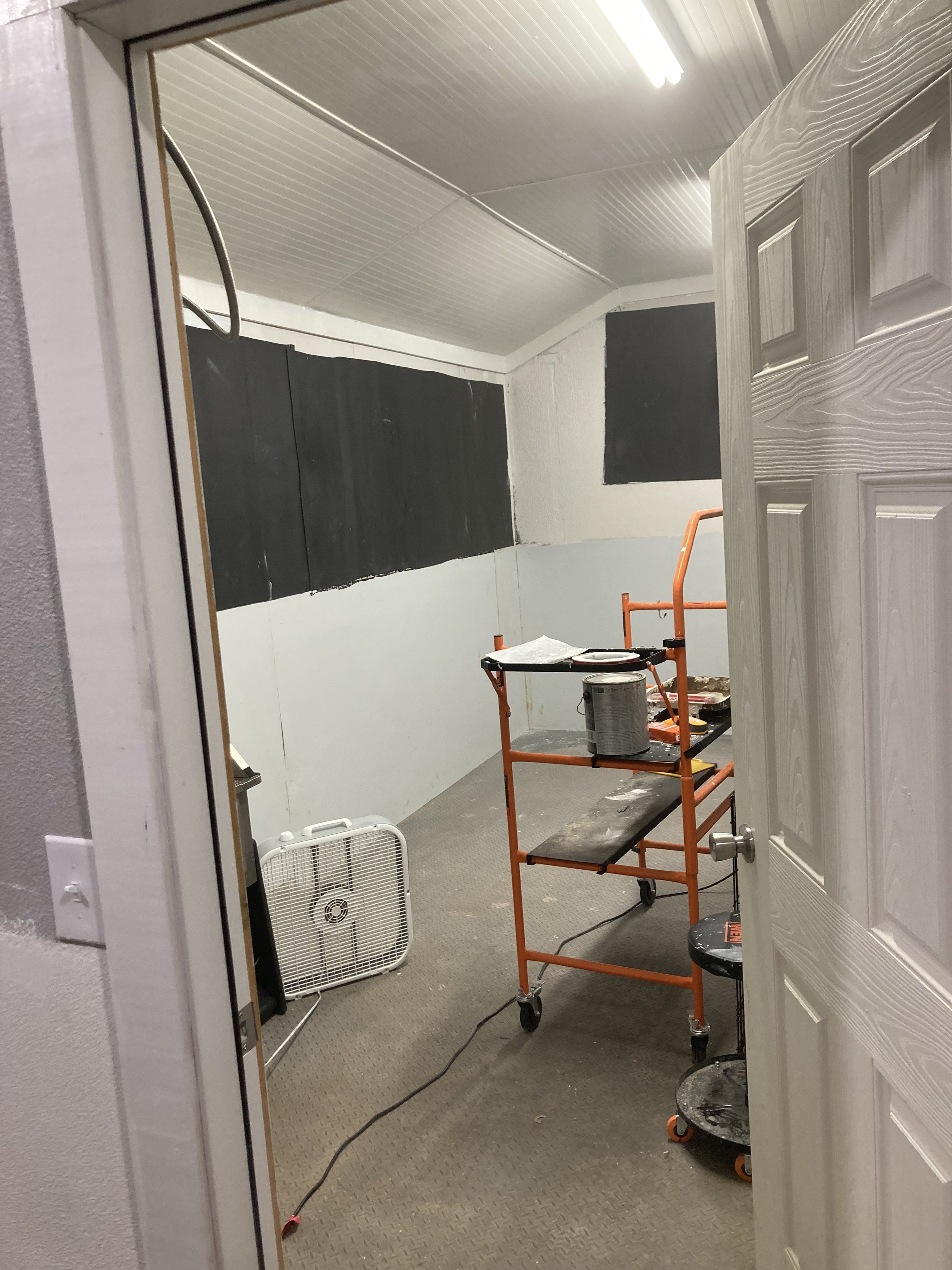The image shows an interior room under construction or renovation with a white ceiling, white walls, and a beige textured floor. There is a small orange scaffold, a black rolling stool, and a white fan. A large portion of a wooden door or panel with a natural wood grain pattern is leaning against the wall. Black rectangular boards are attached to the wall amidst construction materials, and a white fan is placed on the floor near the door frame. An orange extension cord is visible on the floor.