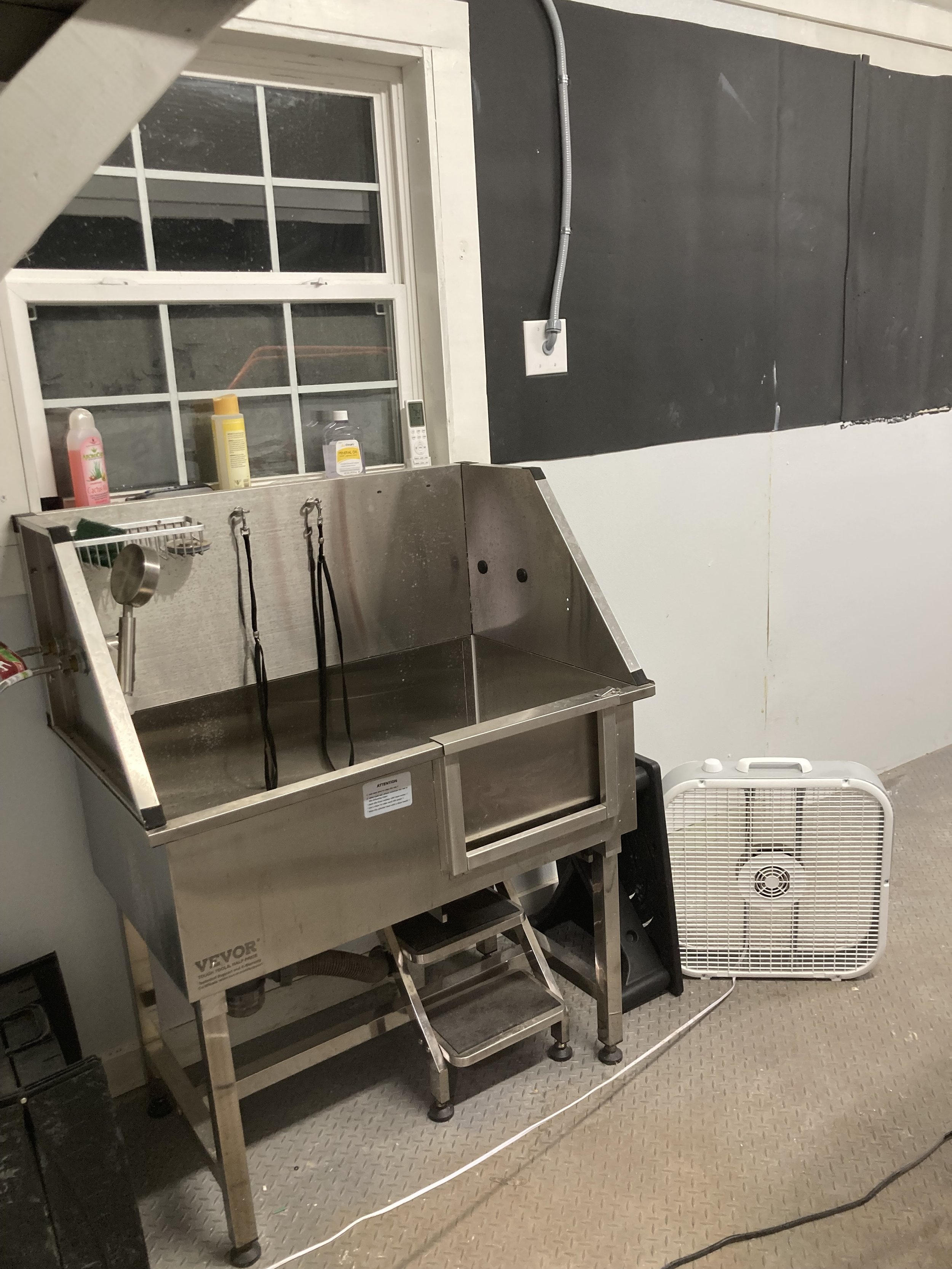 A stainless steel pet washing station with hanging leashes, placed next to a window with cleaning products and a microwave, in a room with black and white walls and an electric fan on the floor.