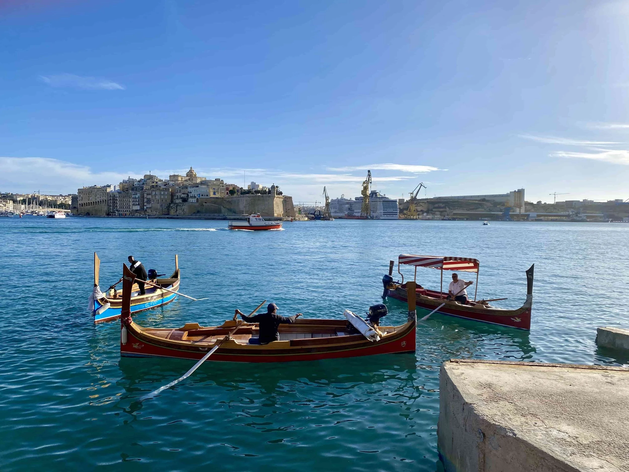 Traditional colorful Luzzu boats docked at the Valletta waterfront with a view of the Three Cities across the Grand Harbour.