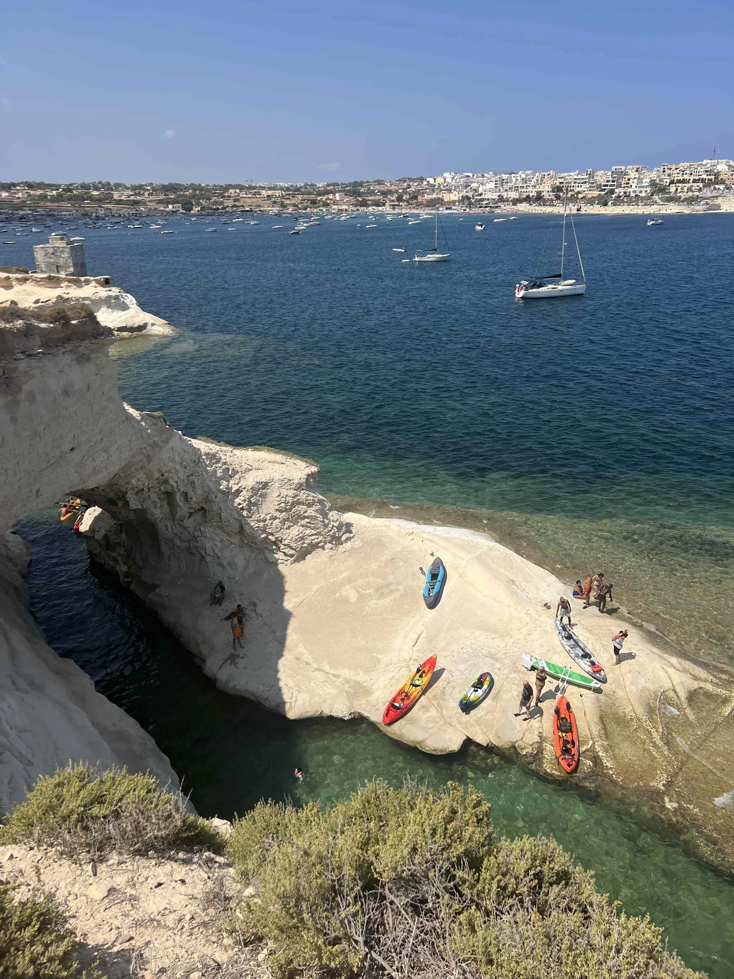 Colourful kayaks resting on rocks near the Munxar Window at St. Thomas Bay in clear turquoise water.