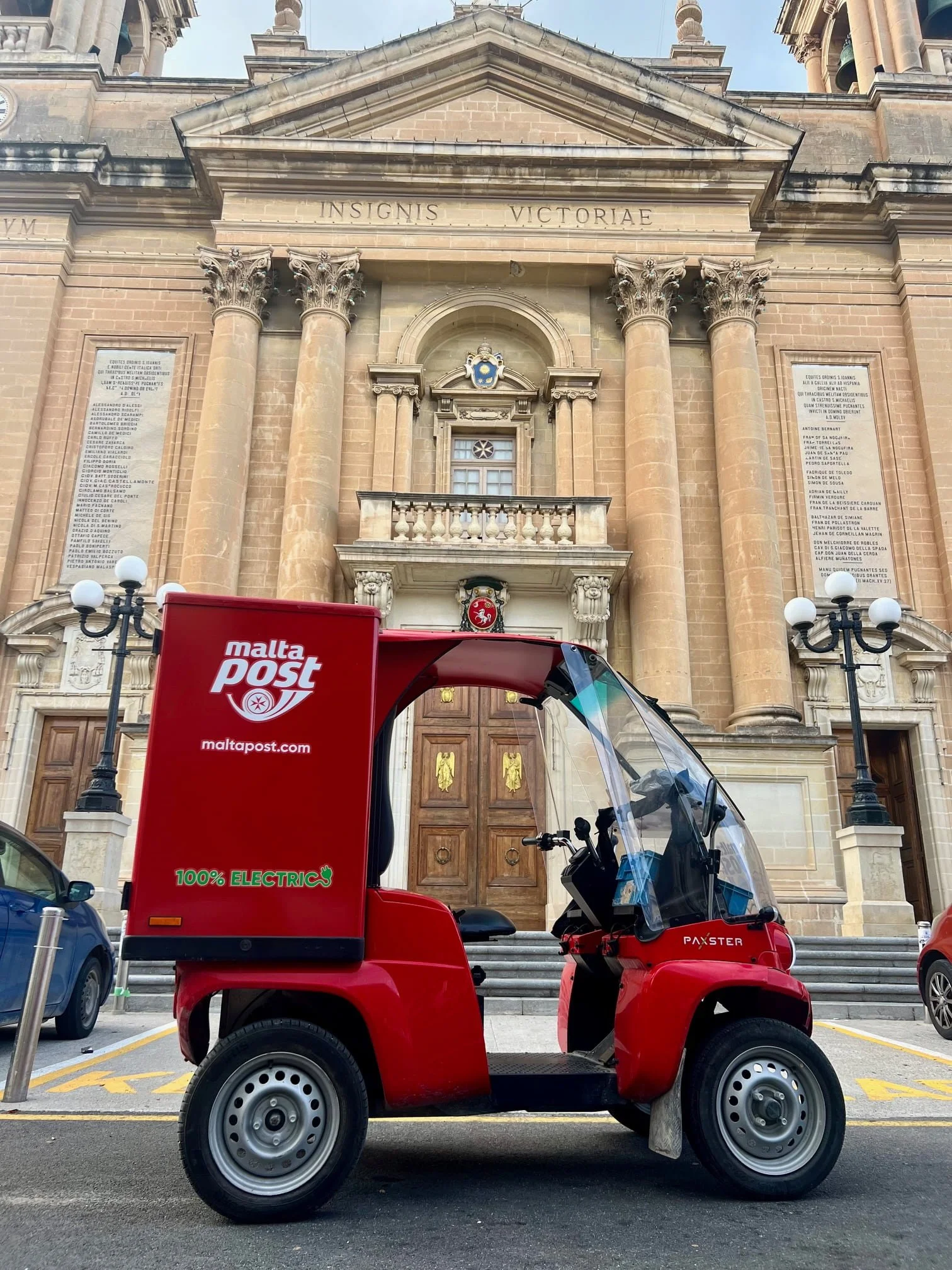 A red electric Maltapost delivery vehicle parked in front of  the Basilica of the Nativity of Mary in Senglea, Malta.