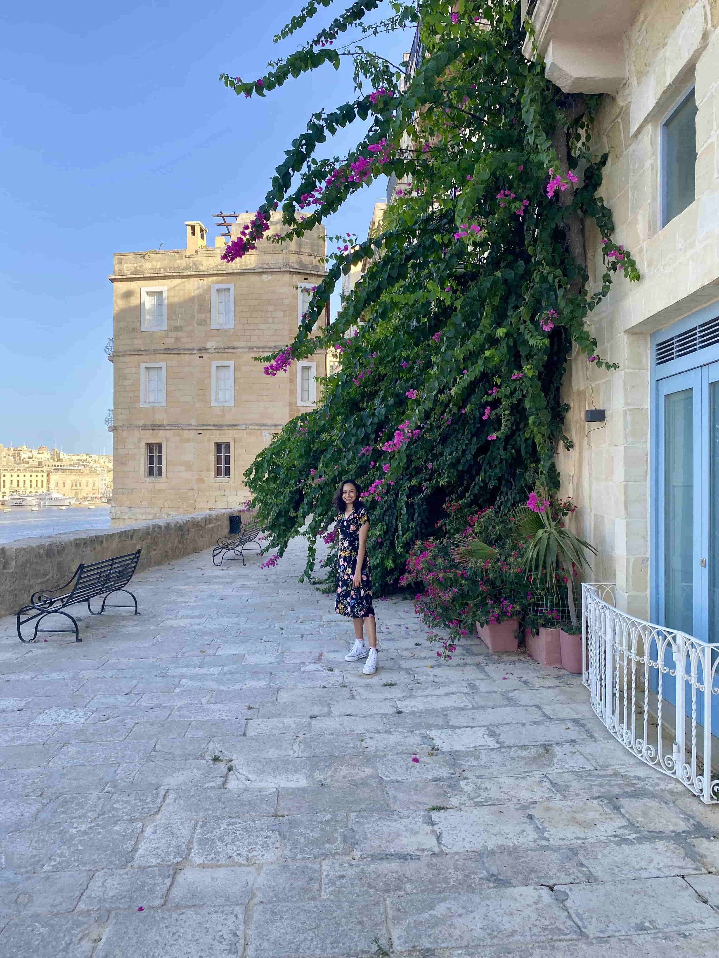 A woman standing next to vibrant purple bougainvillea flowers cascading down a wall near Gardjola Gardens in Senglea.