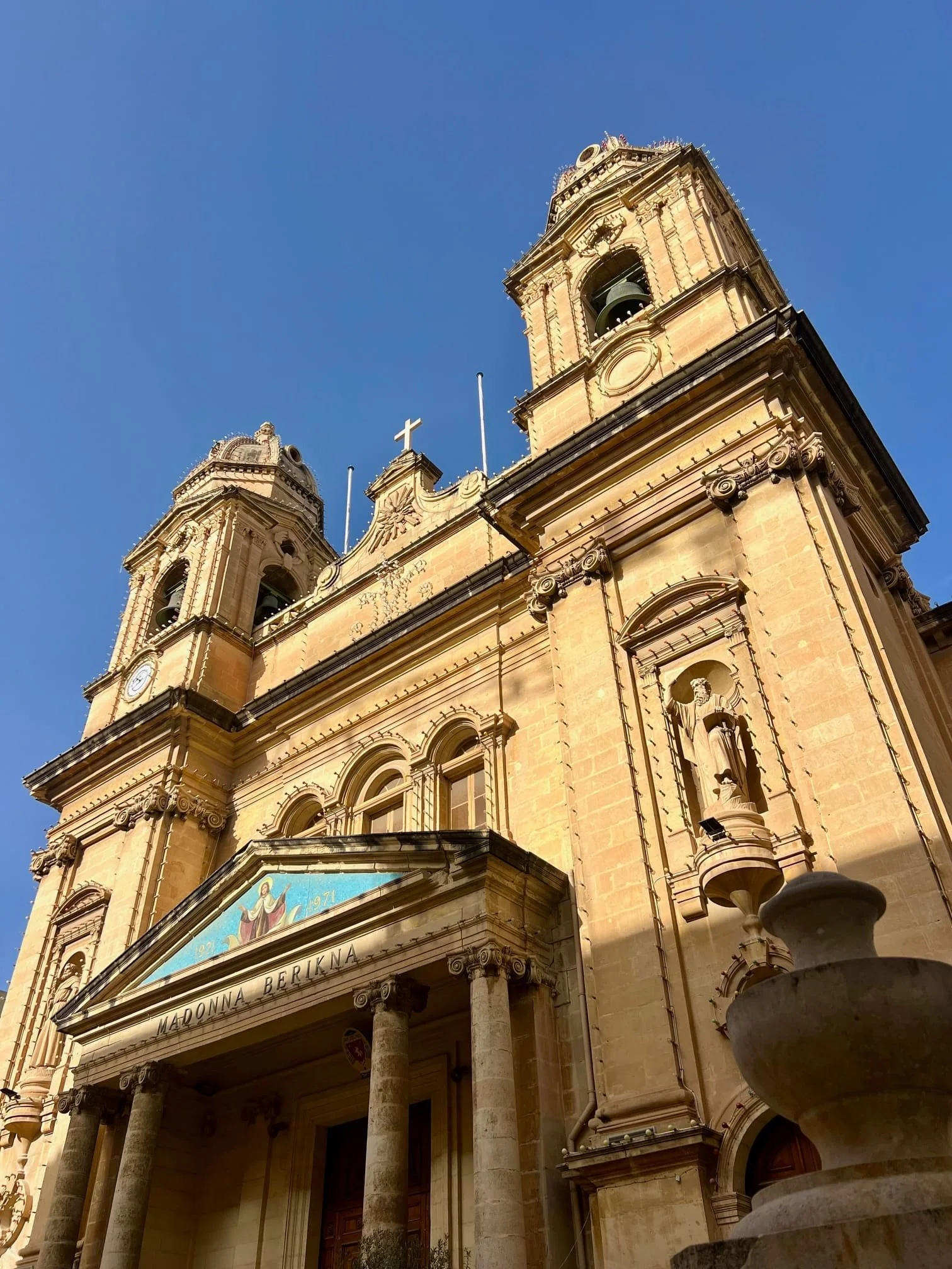 The facade of the Gzira Parish Church (Our Lady of Mount Carmel) with its twin bell towers and golden limestone architecture against a blue sky.