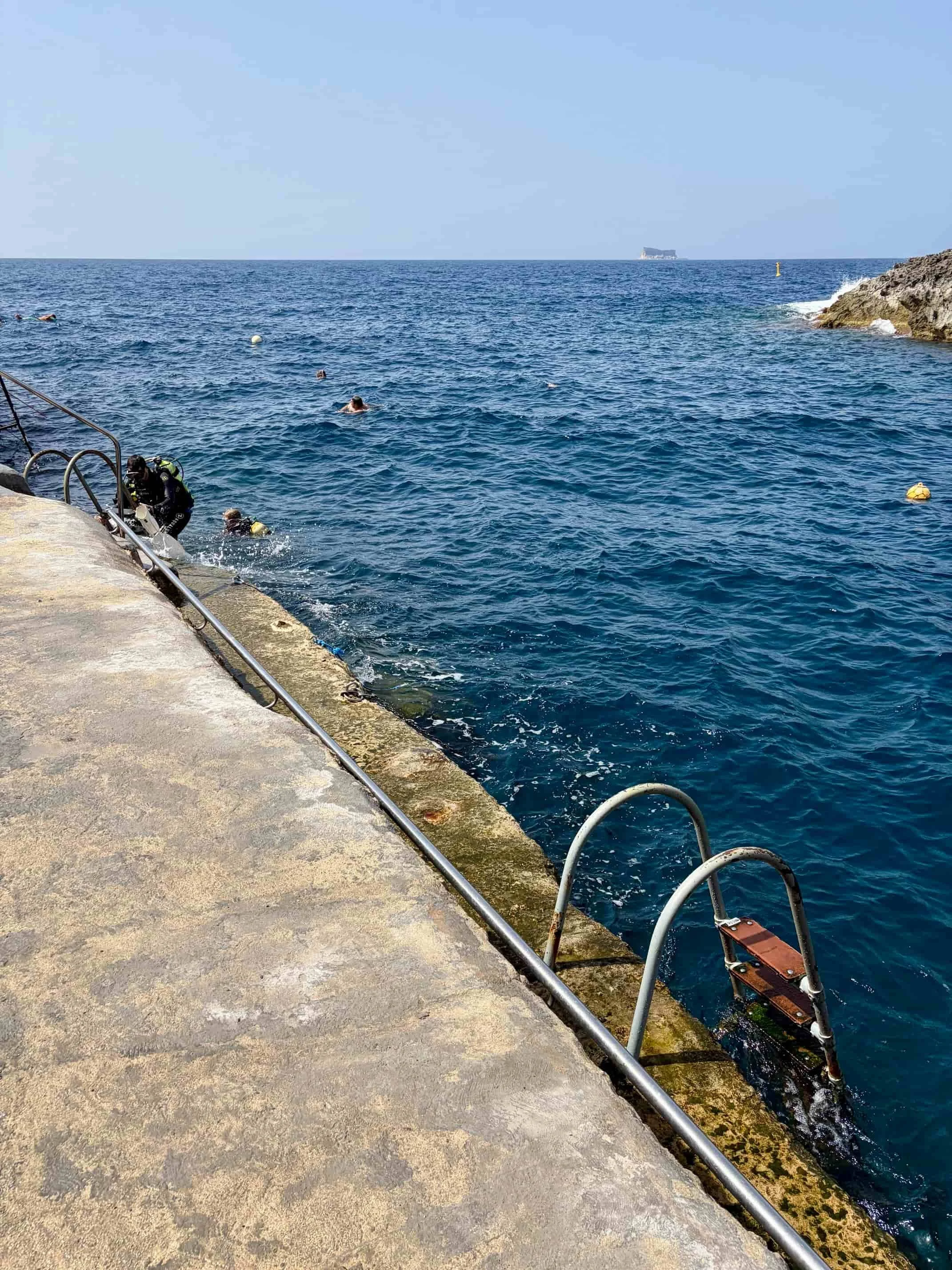 Scuba divers in wetsuits exit the sea via a concrete slipway with a metal handrail at the exit of Wied iz Zurrieq bay, Malta.