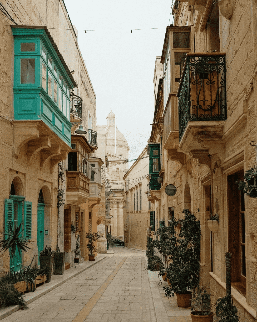 A narrow historic alley in Birgu featuring traditional green wooden balconies and limestone arches.