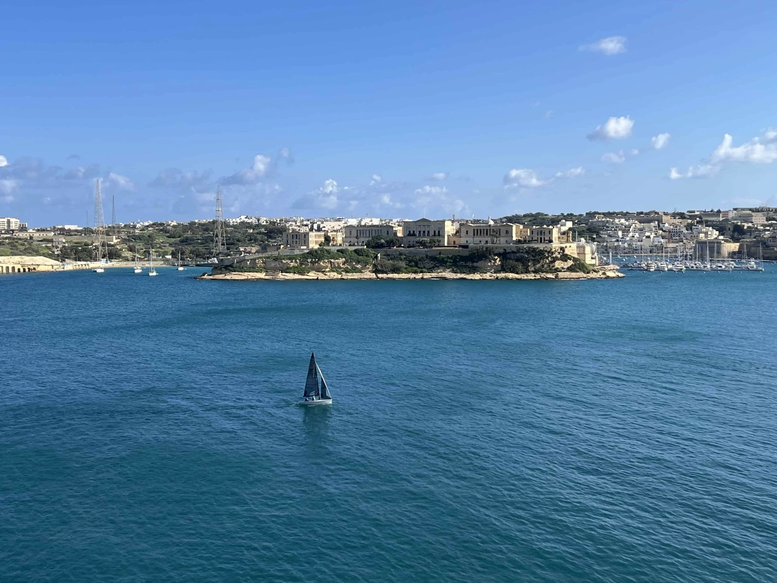 A lone sailing ship navigating the calm waters of the Grand Harbour with the Three Cities in the background.