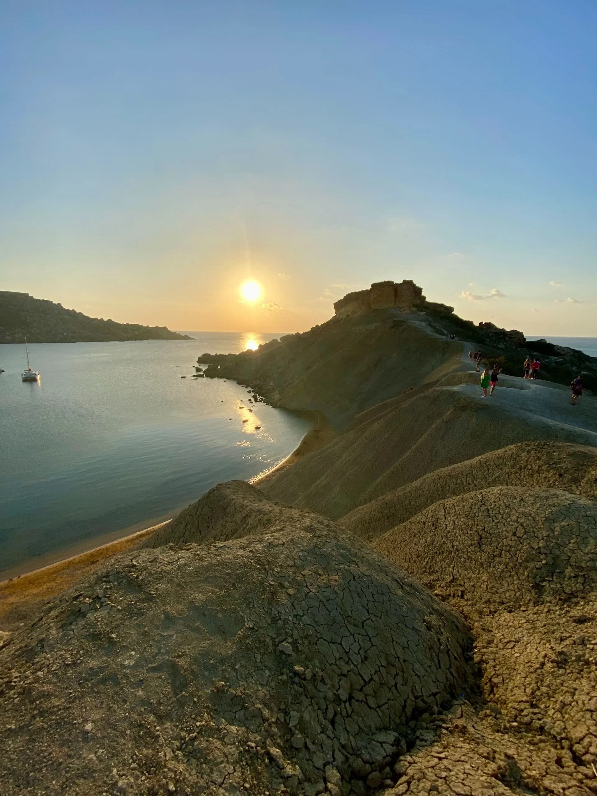 Golden sunset over Gnejna Bay, Malta, illuminating the distinctive clay slopes and a sailboat on the calm water, with hikers walking along the coastal ridge.
