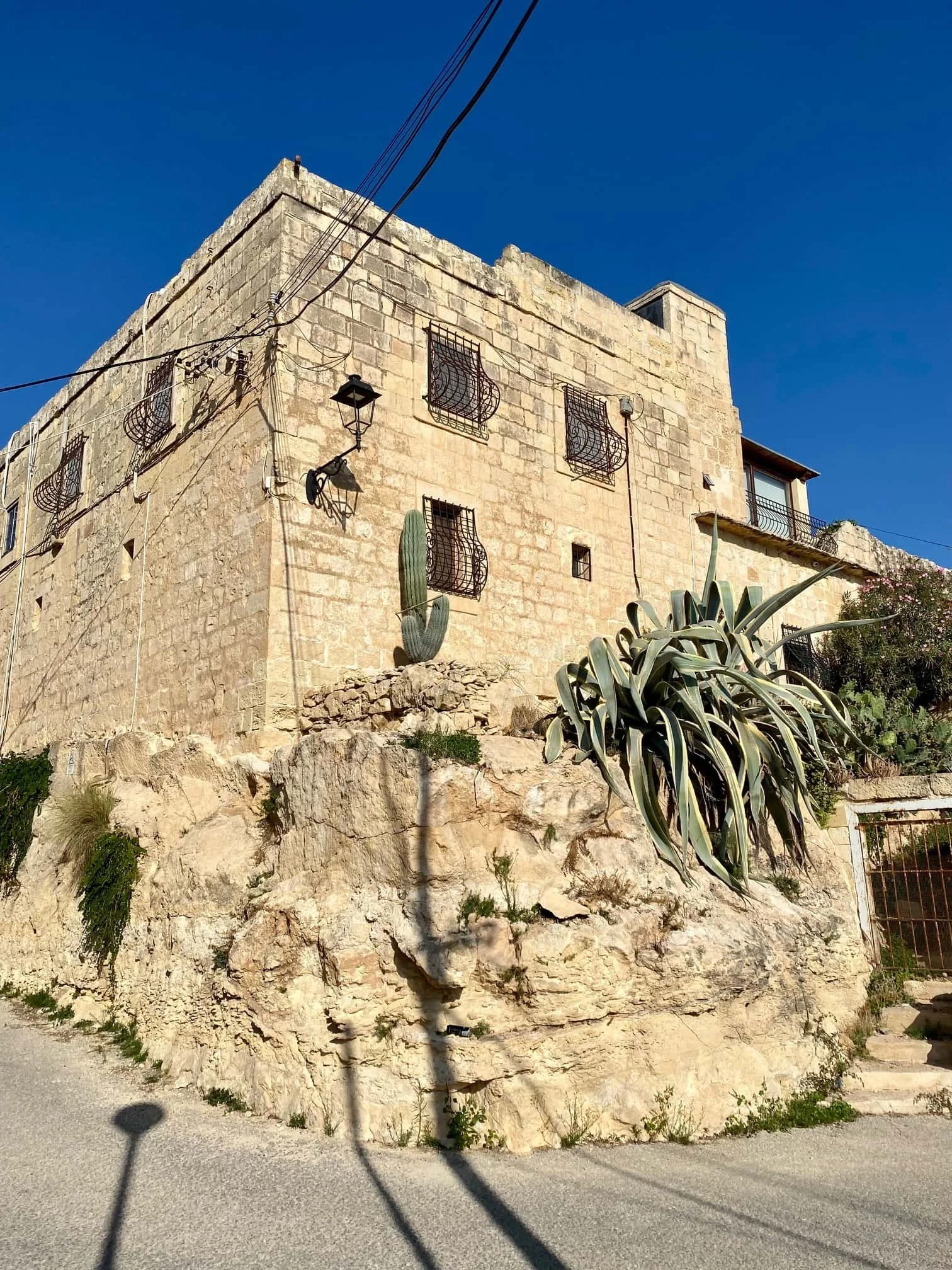 A traditional Maltese stone townhouse built on a rocky incline in Marsaskala, featuring old wooden windows and a large cactus plant in the foreground.