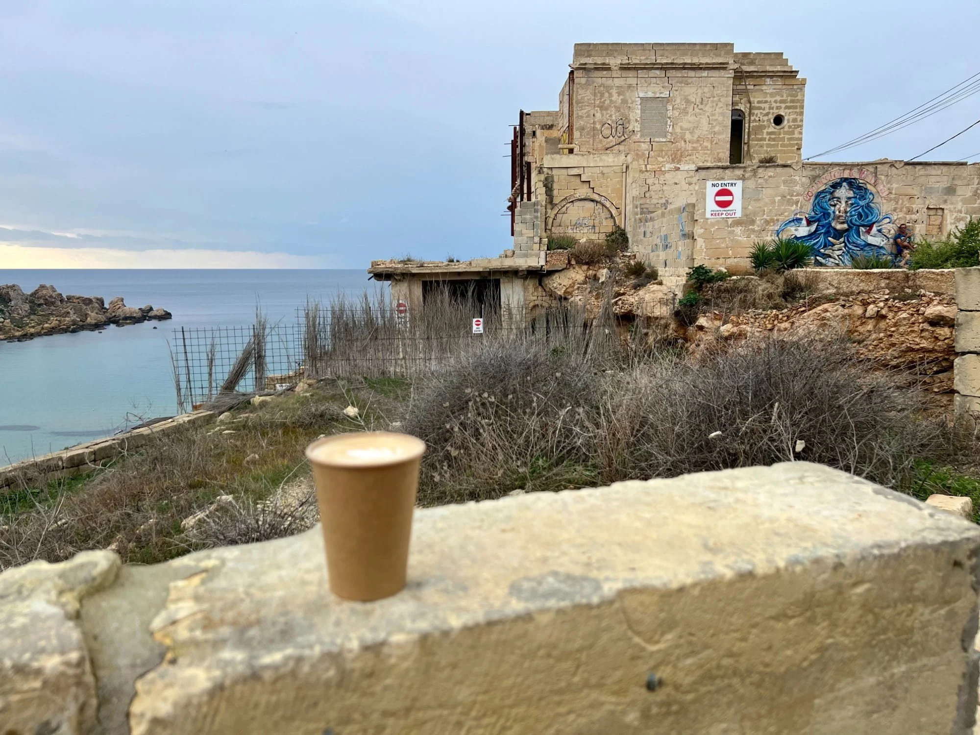 A coffee cup on a wall and an older building with mural in the background.