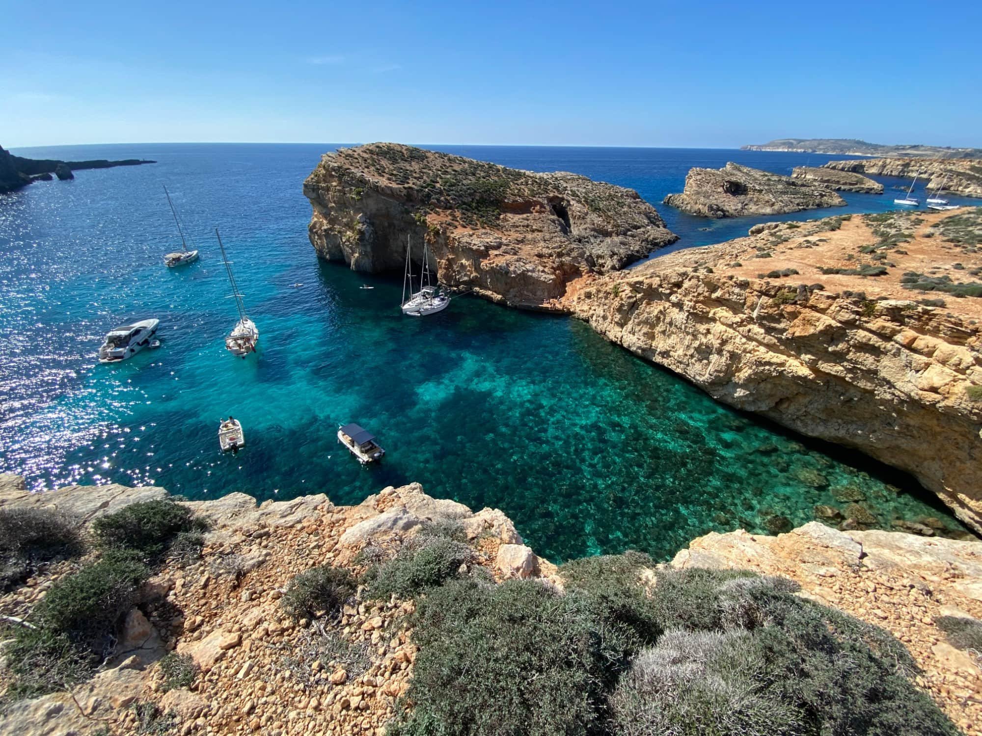 A high-angle panoramic view of the inlets and bays of Comino Island filled with anchored boats.