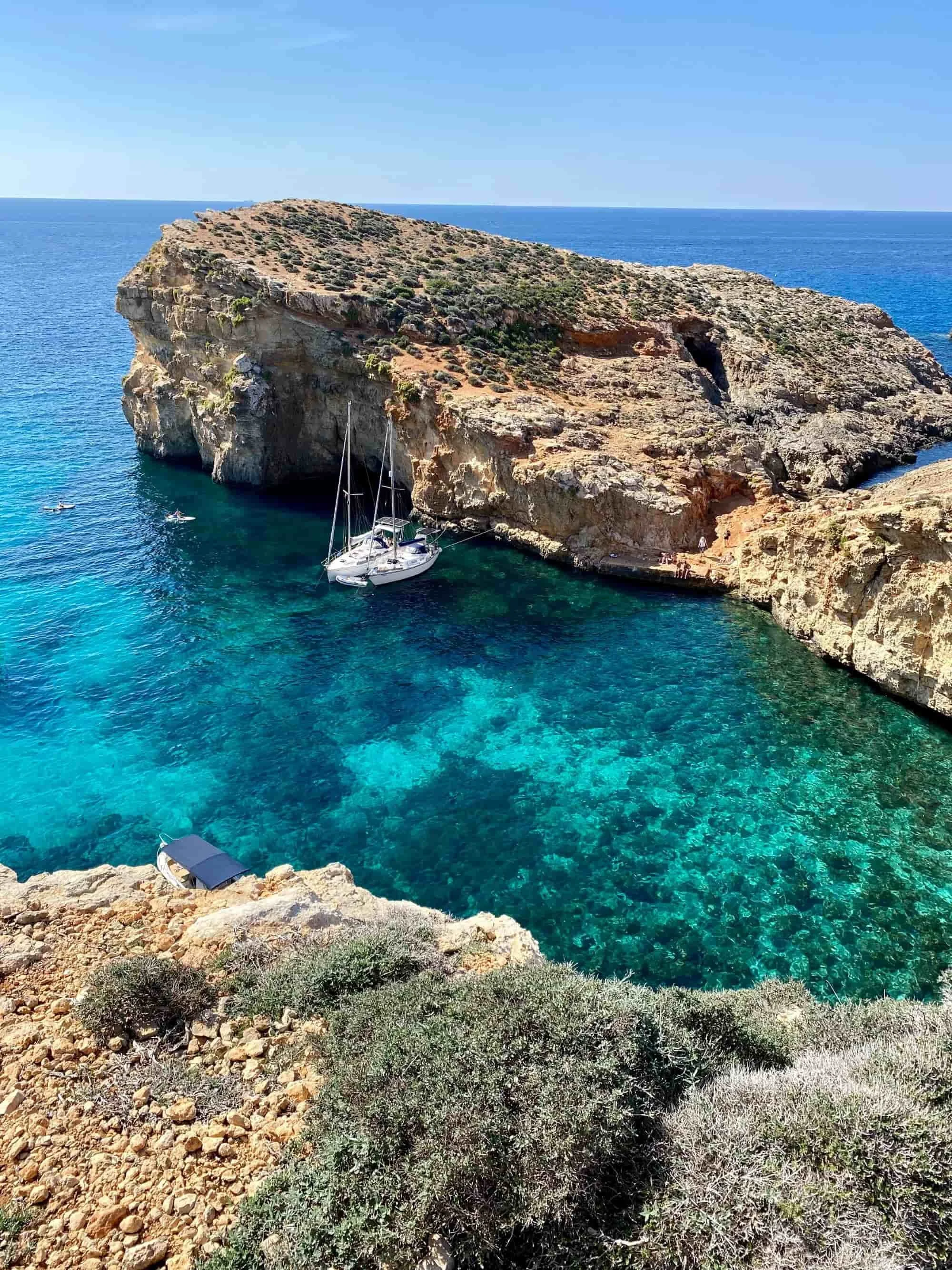 Rugged limestone cliffs and sea caves on Comino Island surrounded by deep teal Mediterranean water.