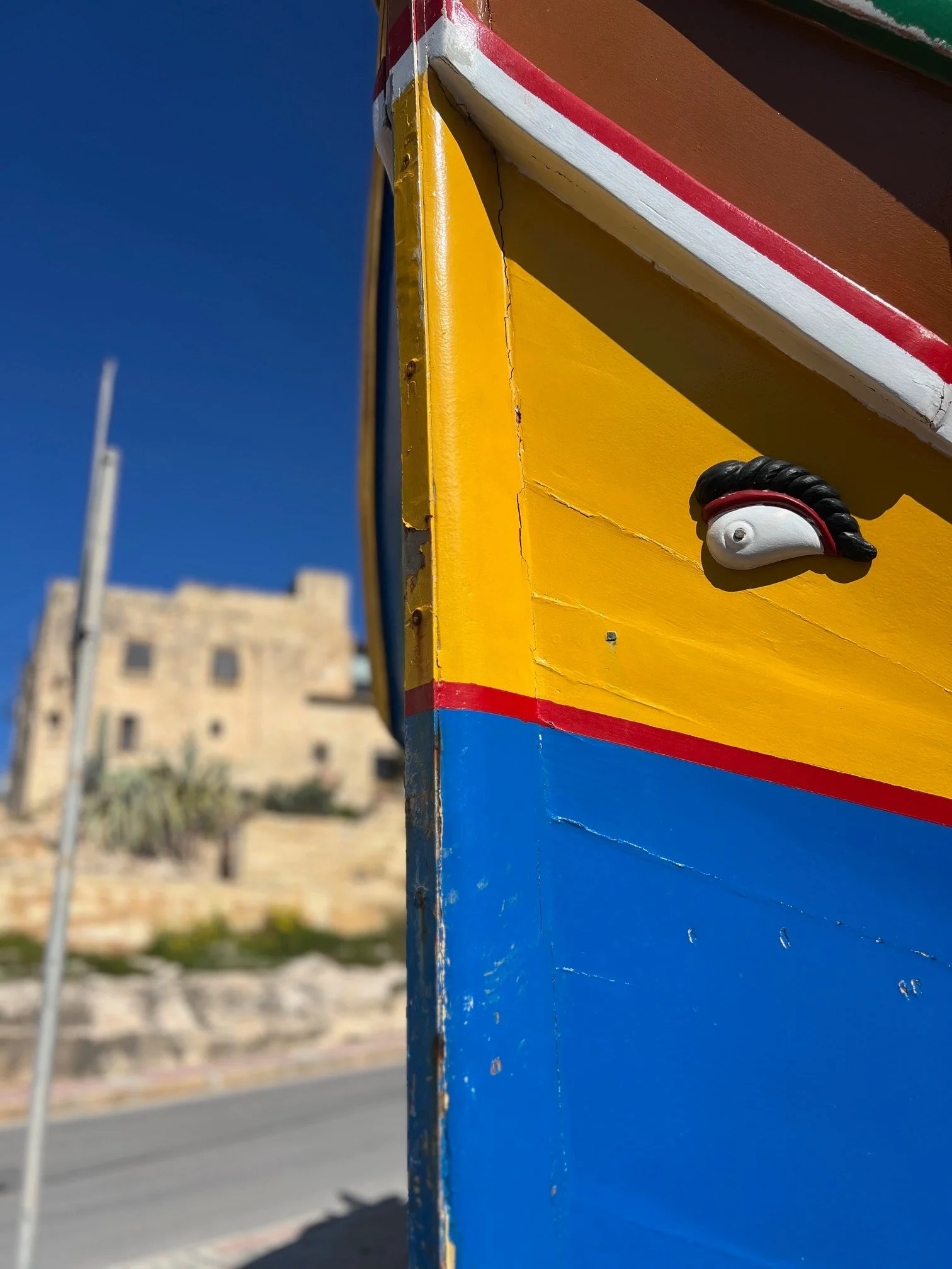 Close-up of a colorful traditional Maltese Luzzu fishing boat with the 'Eye of Horus' symbol, with an old stone townhouse in Marsaskala visible in the background.