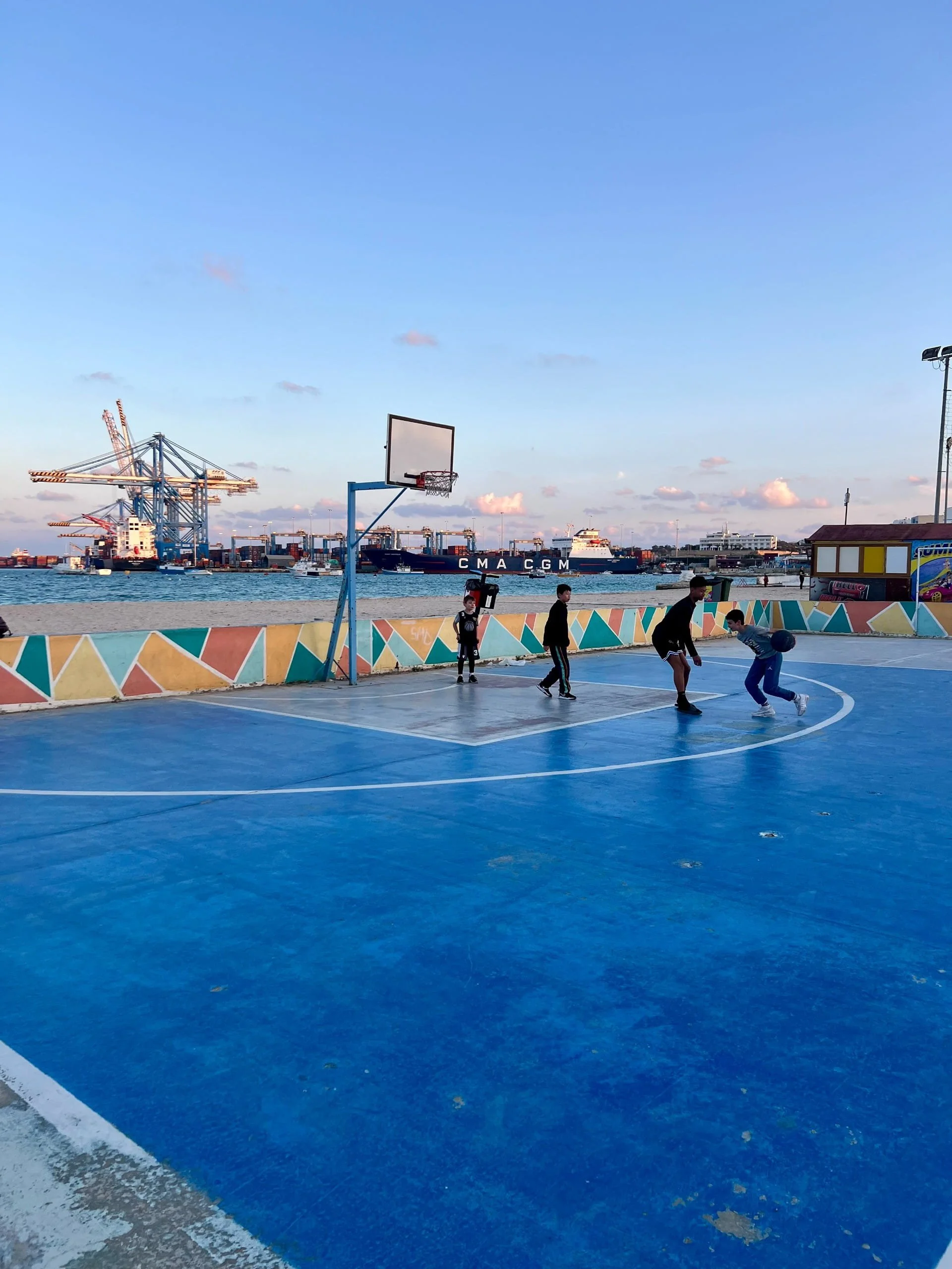 The basketball court at Pretty Bay illuminated by soft sunset light with port cranes in the distance.