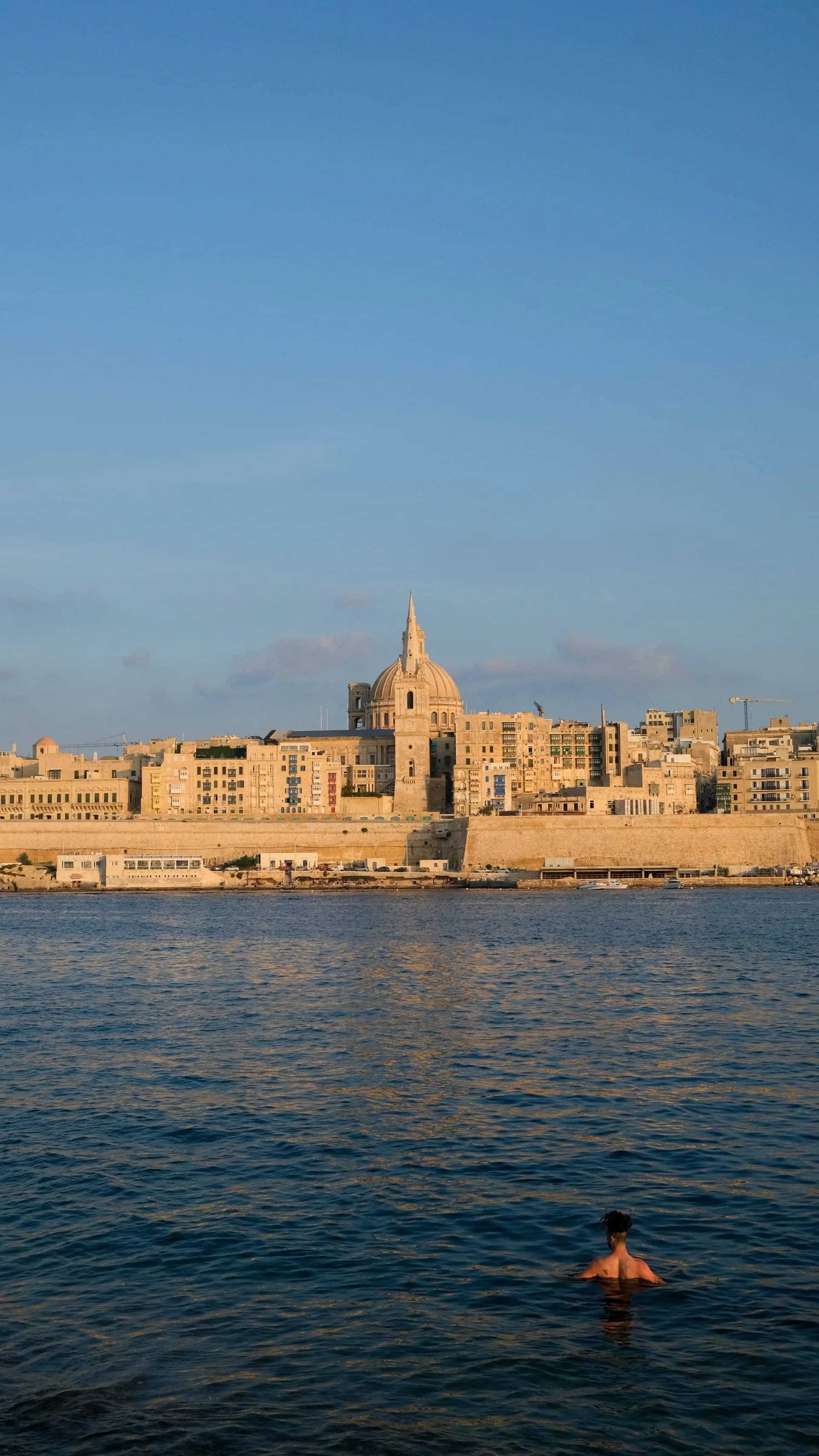 Rocky coastline of Stone Beach on Manoel Island looking across the blue water towards the Valletta fortifications.