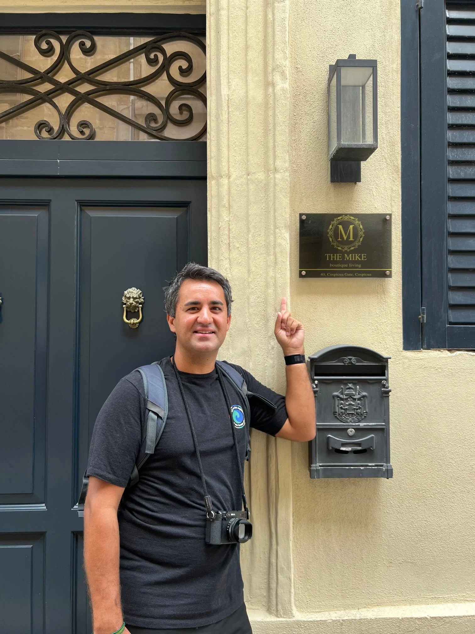 Mike from the ExplorinMalta team standing in front of 'The Mike' hotel in Valletta, smiling and pointing at the name plaque.