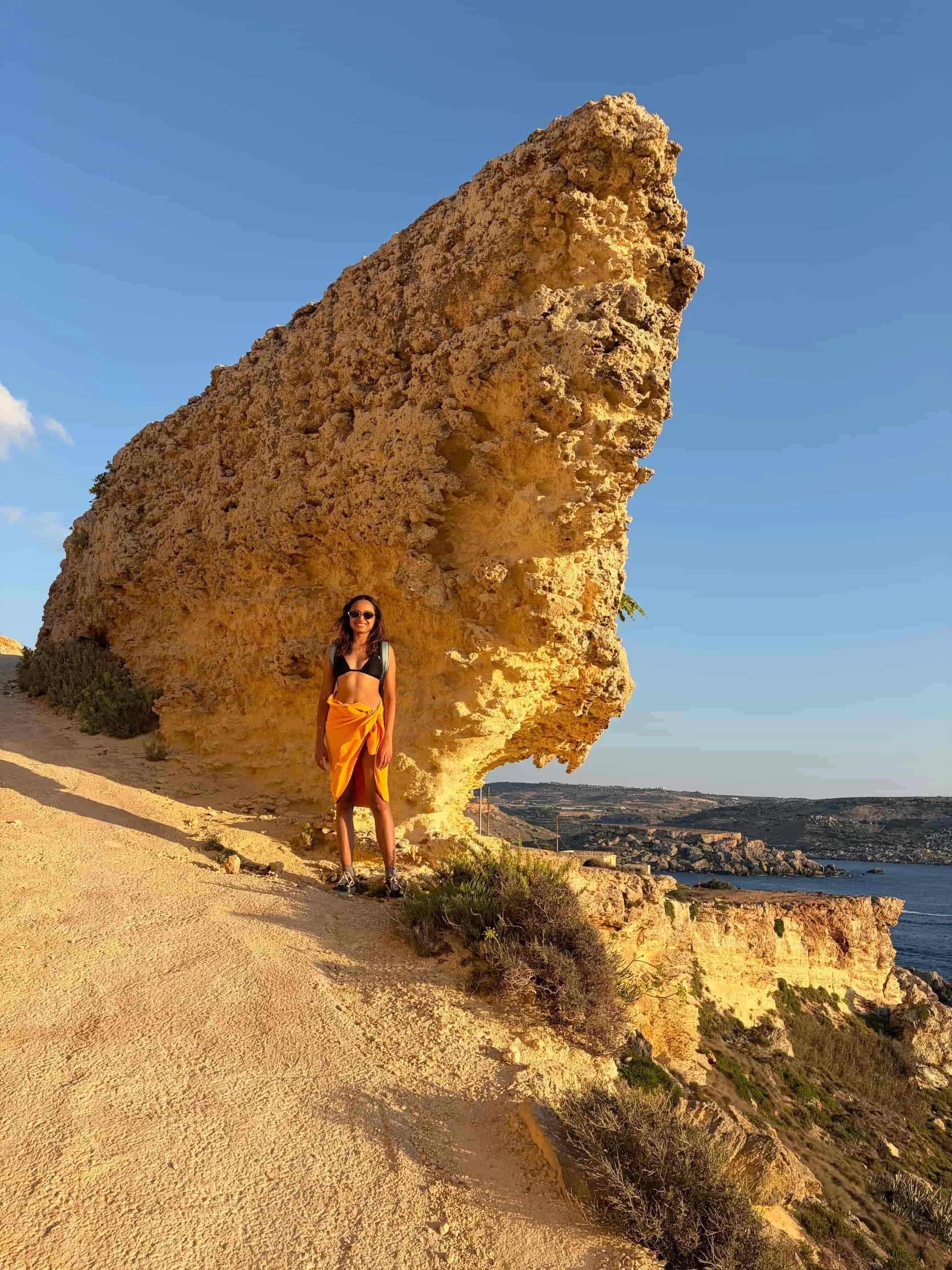 Person standing next to a huge, wind-eroded limestone rock formation on the clifftop path near Ras il-Wahx, Malta.