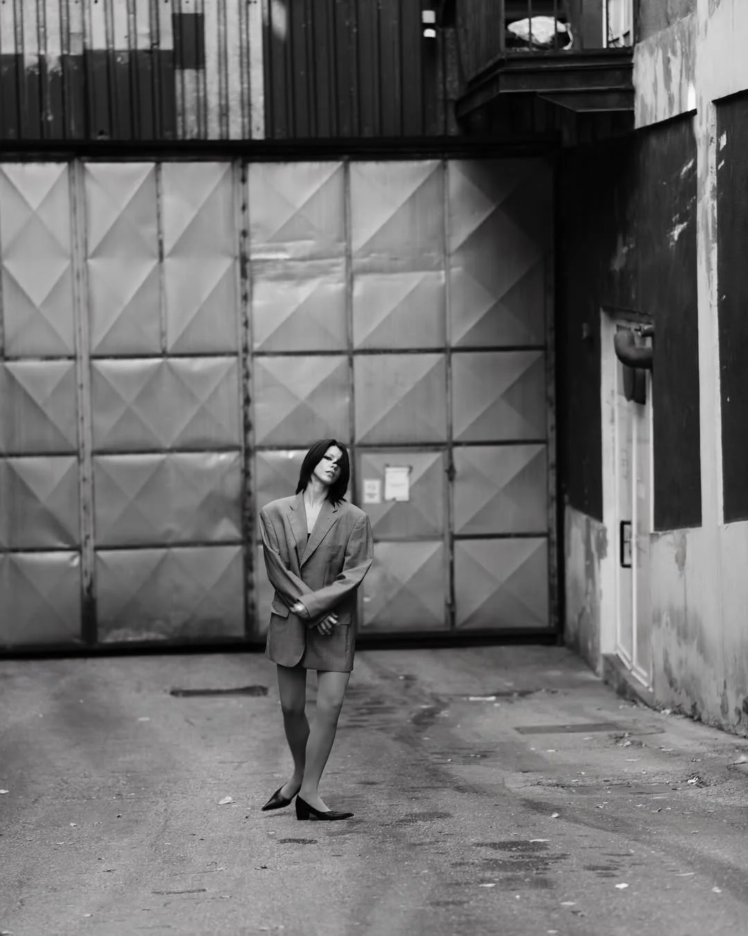 Black and white photo of a person wearing an oversized blazer, standing in front of a large metal garage door. The setting is industrial, with visible concrete and metal textures.
