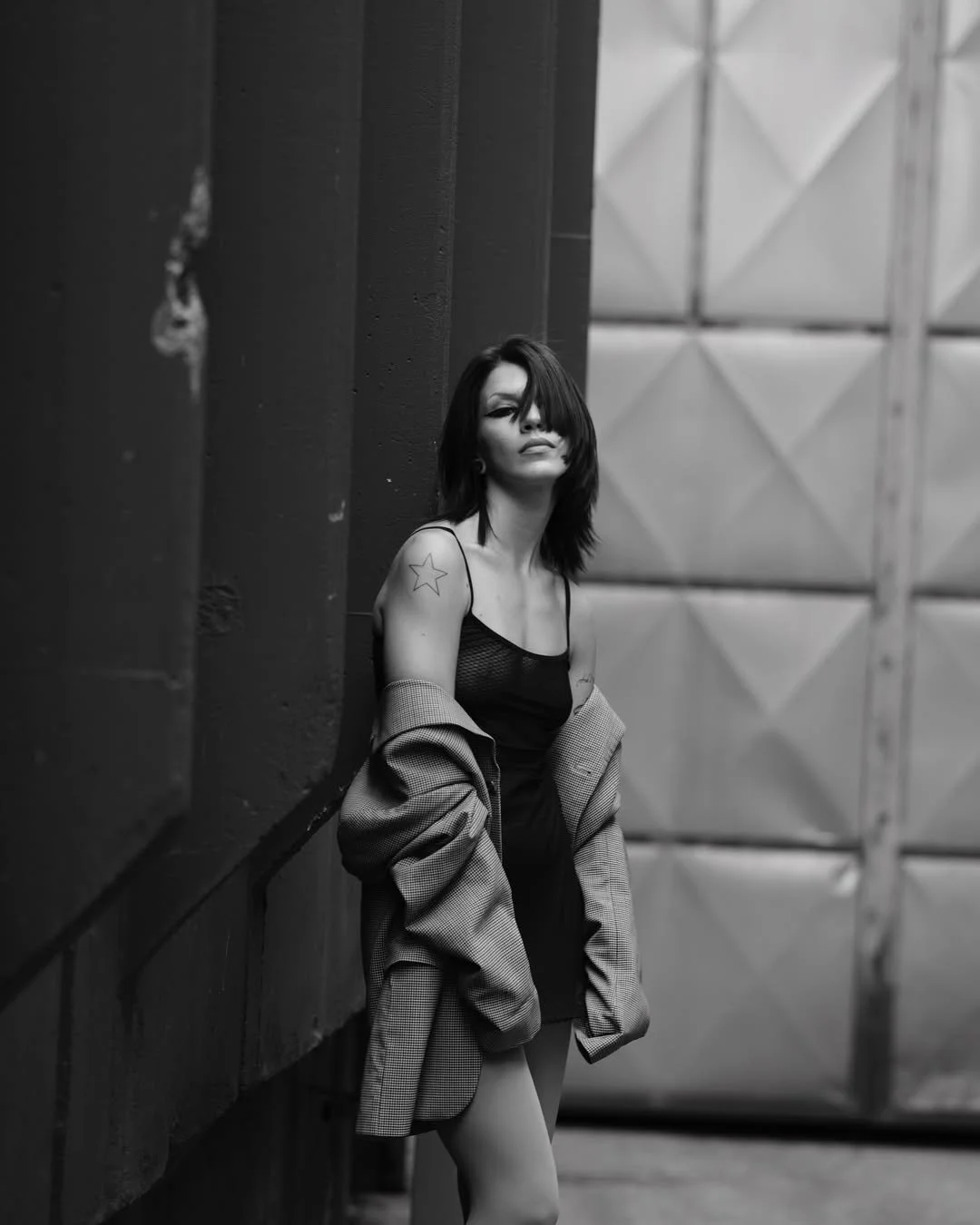 Black and white photo of a woman with a star tattoo on her arm wearing a dress and jacket, leaning against a wall in an urban setting.