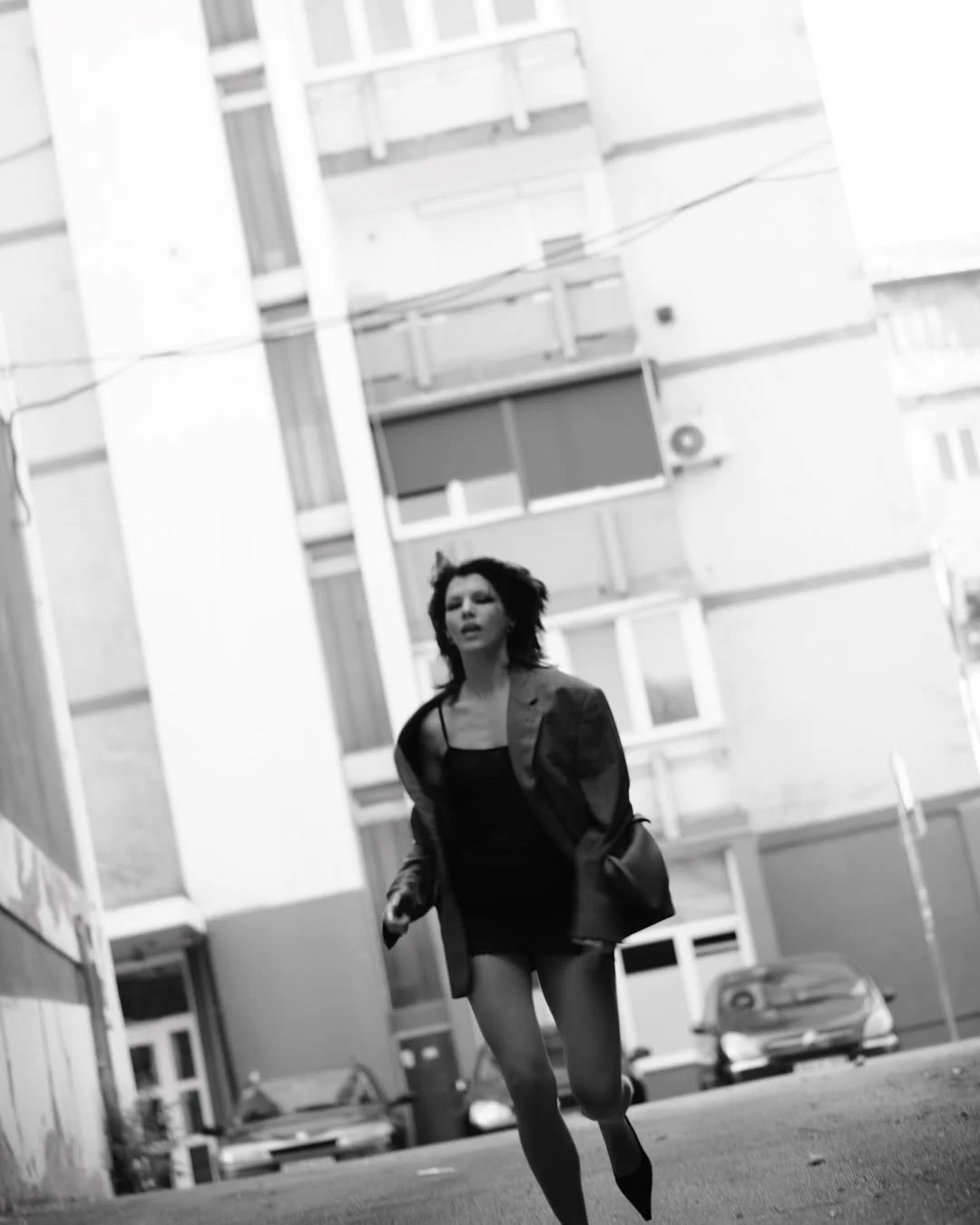 Black and white photo of a person in a blazer and dress running in an urban alley with tall buildings in the background.