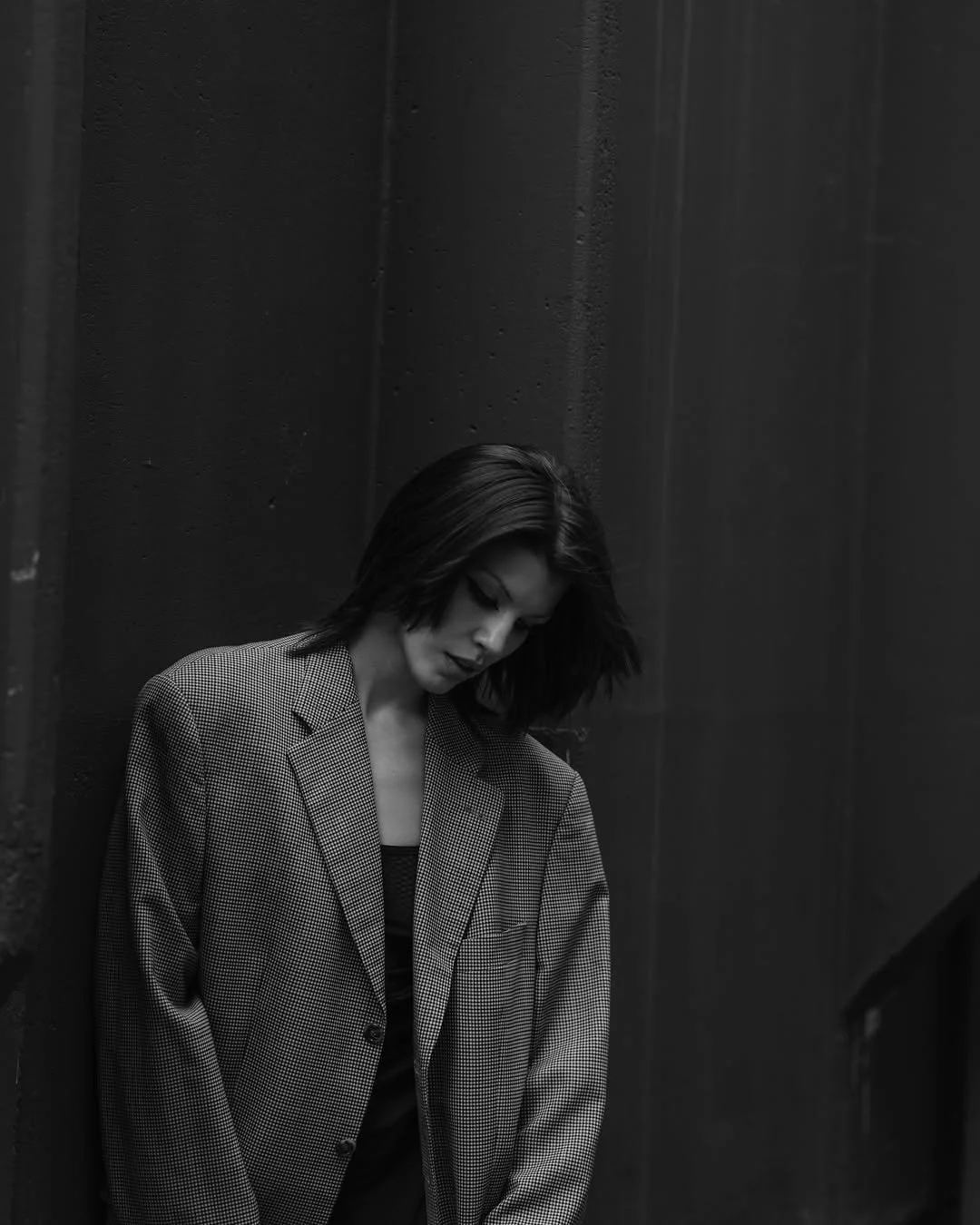 Black and white photo of a person leaning against a wall, wearing an oversized blazer, with head tilted downward.