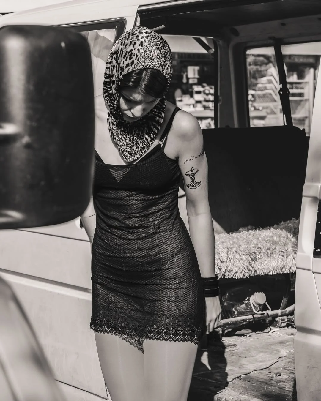 Black and white photo of a person in a lace dress and leopard print headscarf standing outside a vehicle with an open door.
