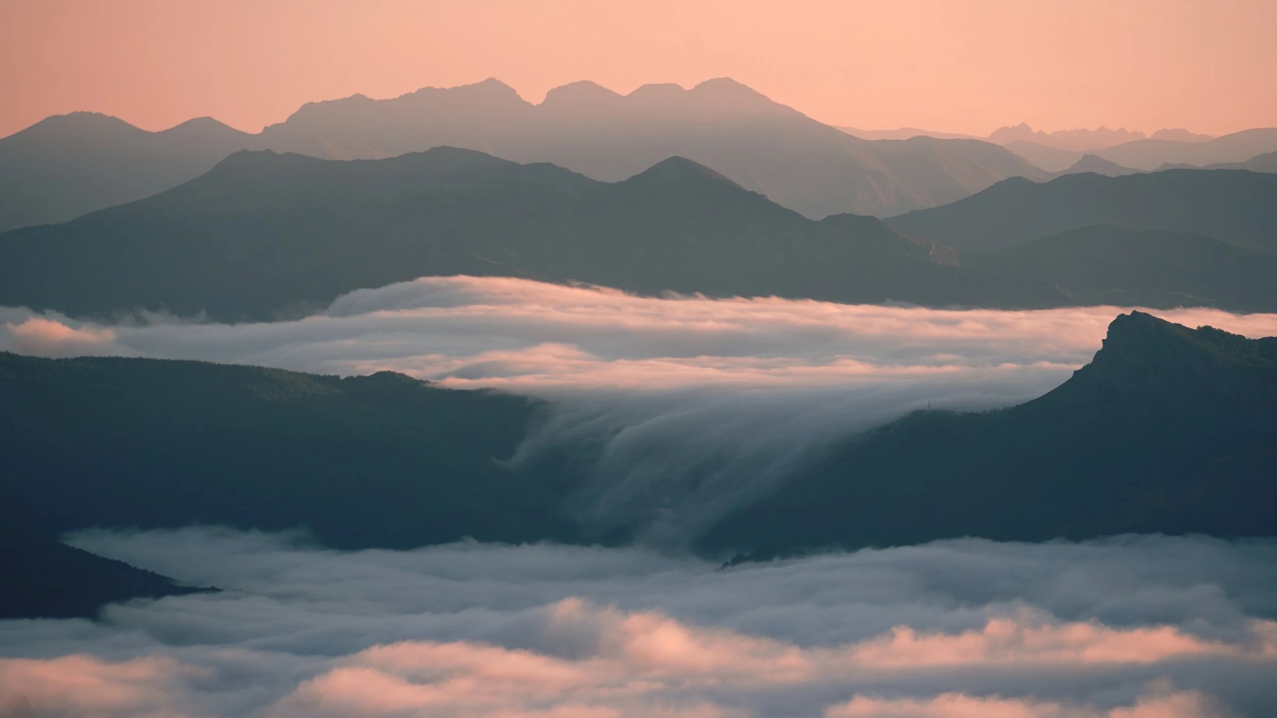 Layers of mountain ranges at sunrise with clouds filling the valleys, pink and orange sky.