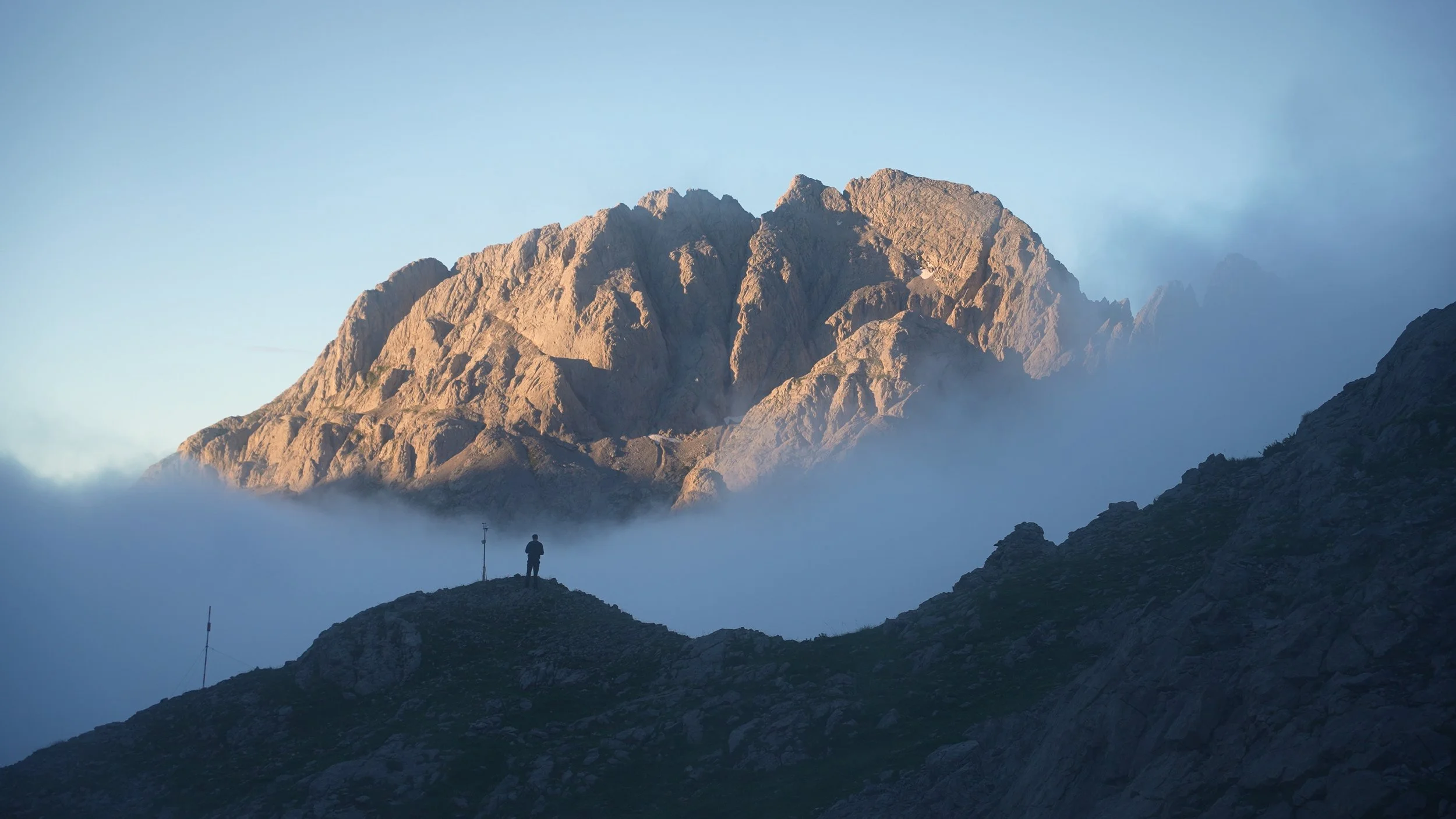 A person standing on a rocky hillside looking at a large mountain range in the distance, with low-hanging clouds surrounding the hills and mountains.