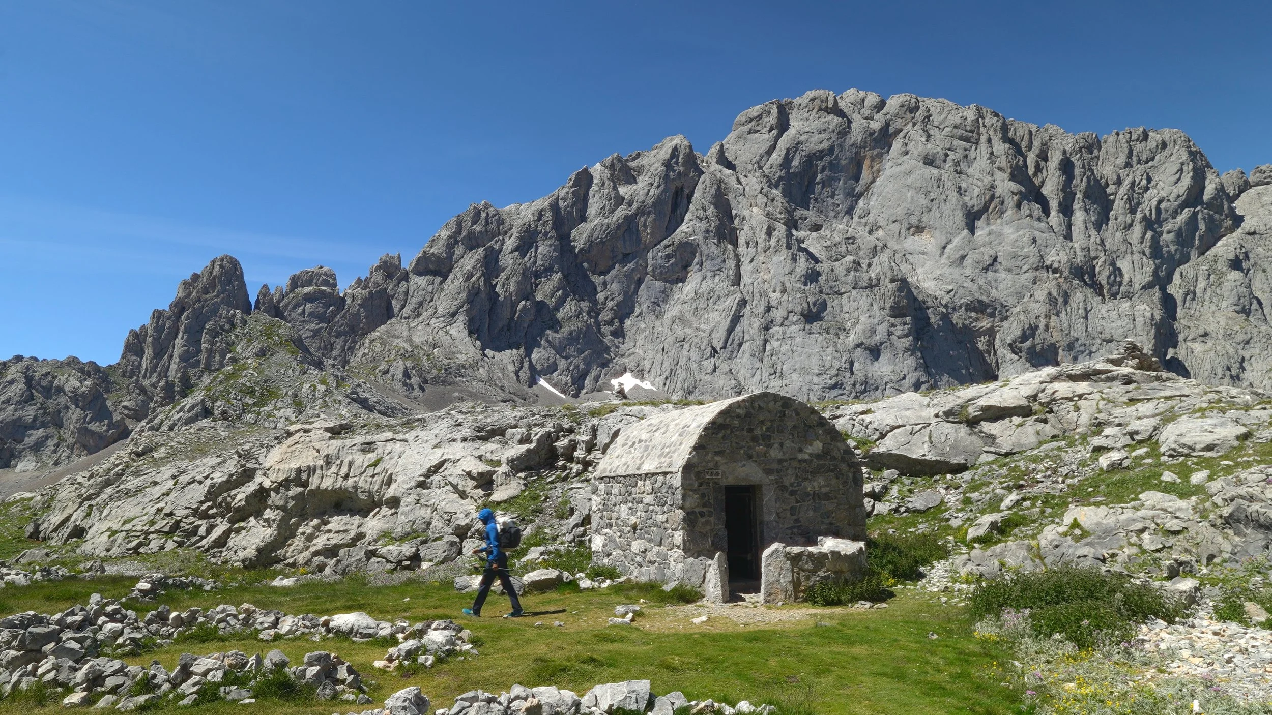 A hiker walking past a small stone shelter in a mountainous landscape with rocky peaks and patches of snow under a clear blue sky.