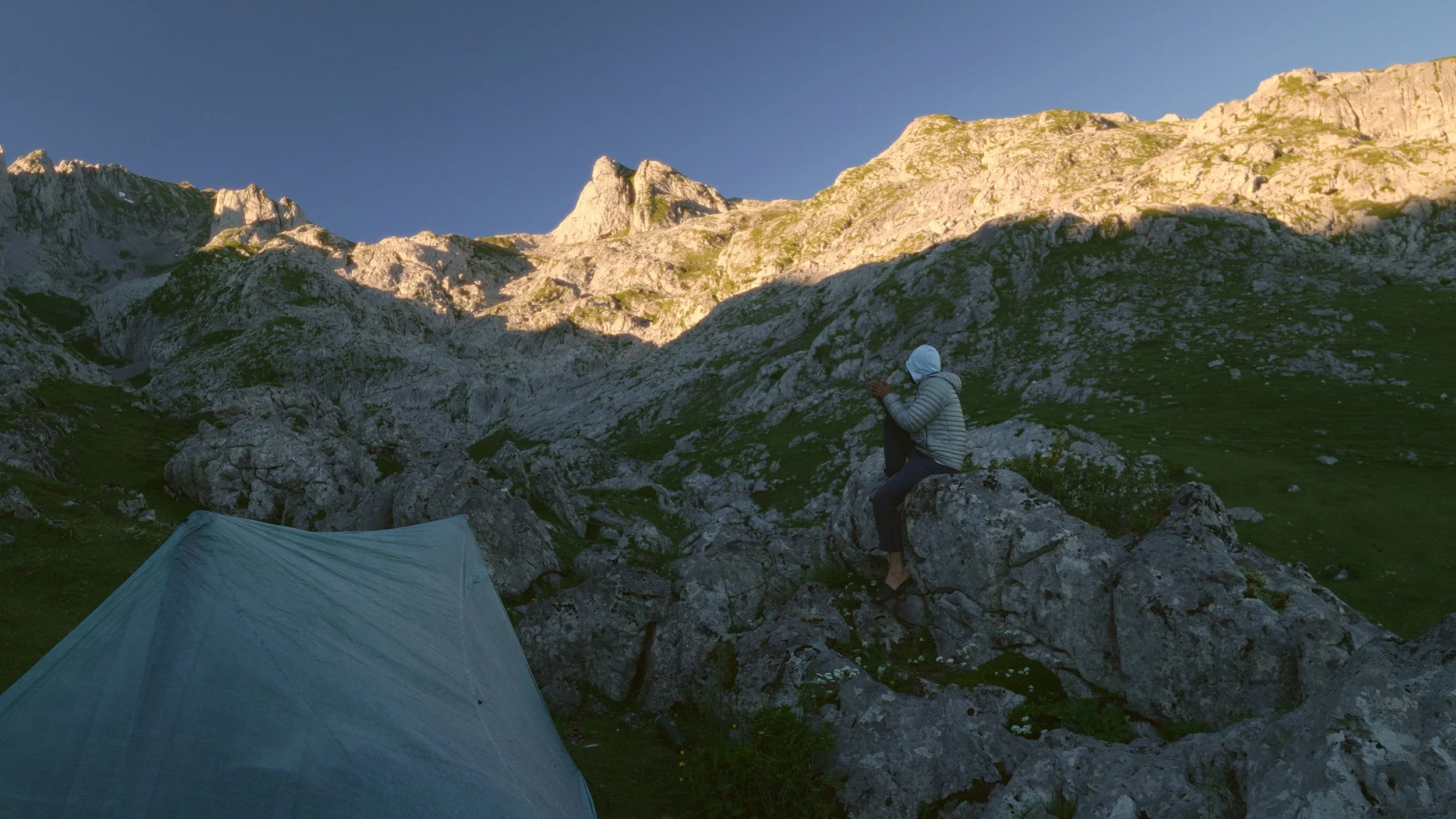 A person sitting on a rock in a mountainous area during daylight, with a tent in the foreground and rugged cliffs in the background.