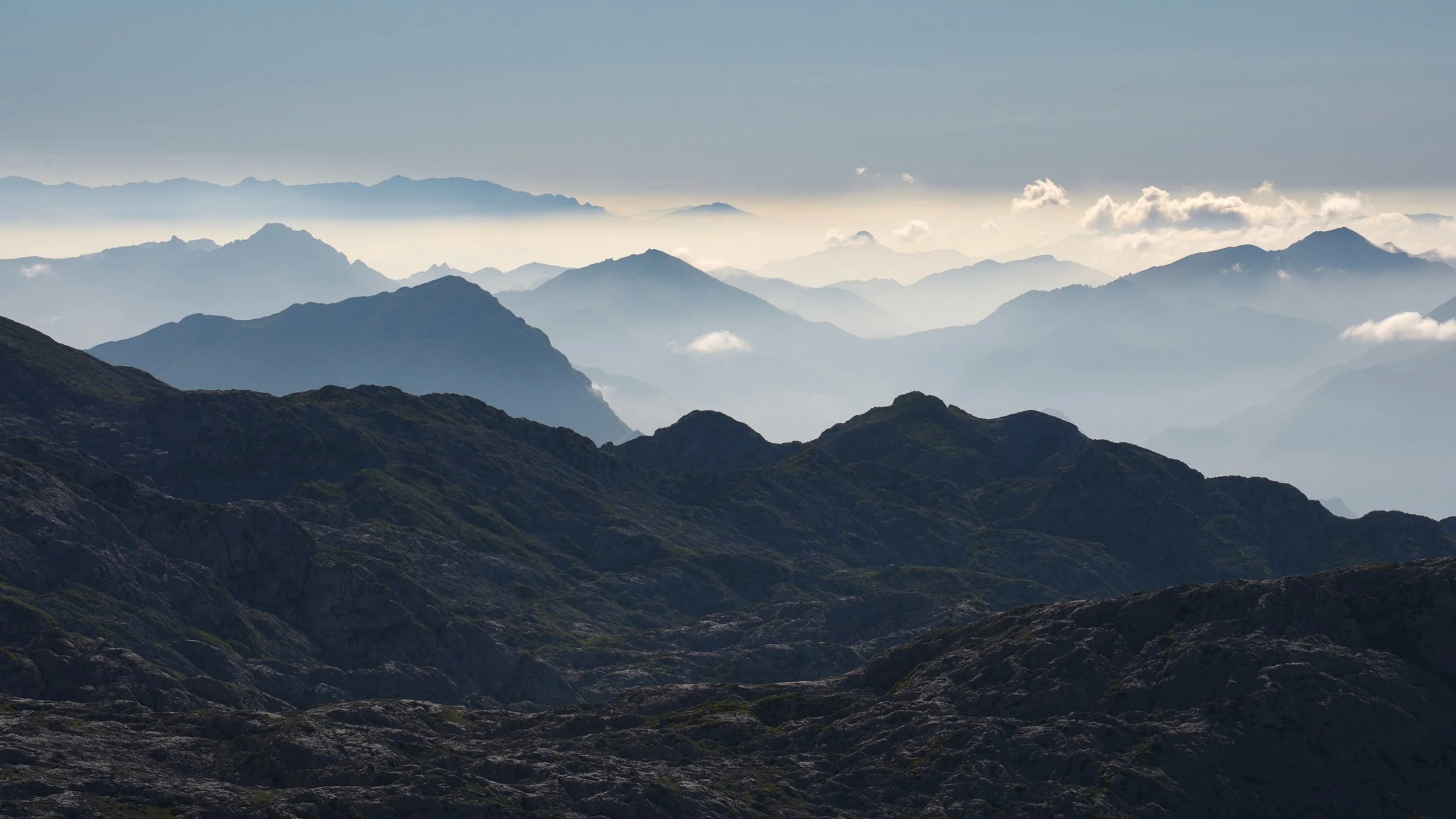 A mountain landscape with multiple layers of ridges and peaks, some covered in clouds, with a hazy sky in the background.
