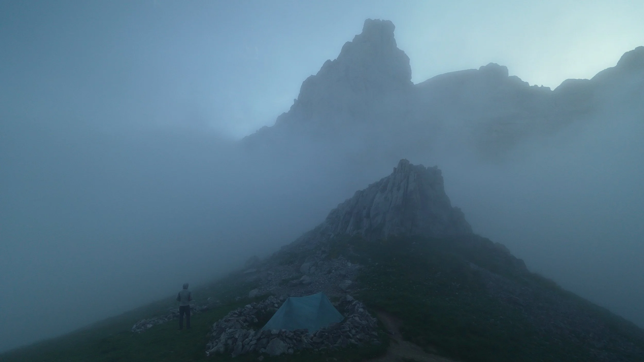 A person standing near a blue camping tent on a grassy hill, with foggy mountains in the background.