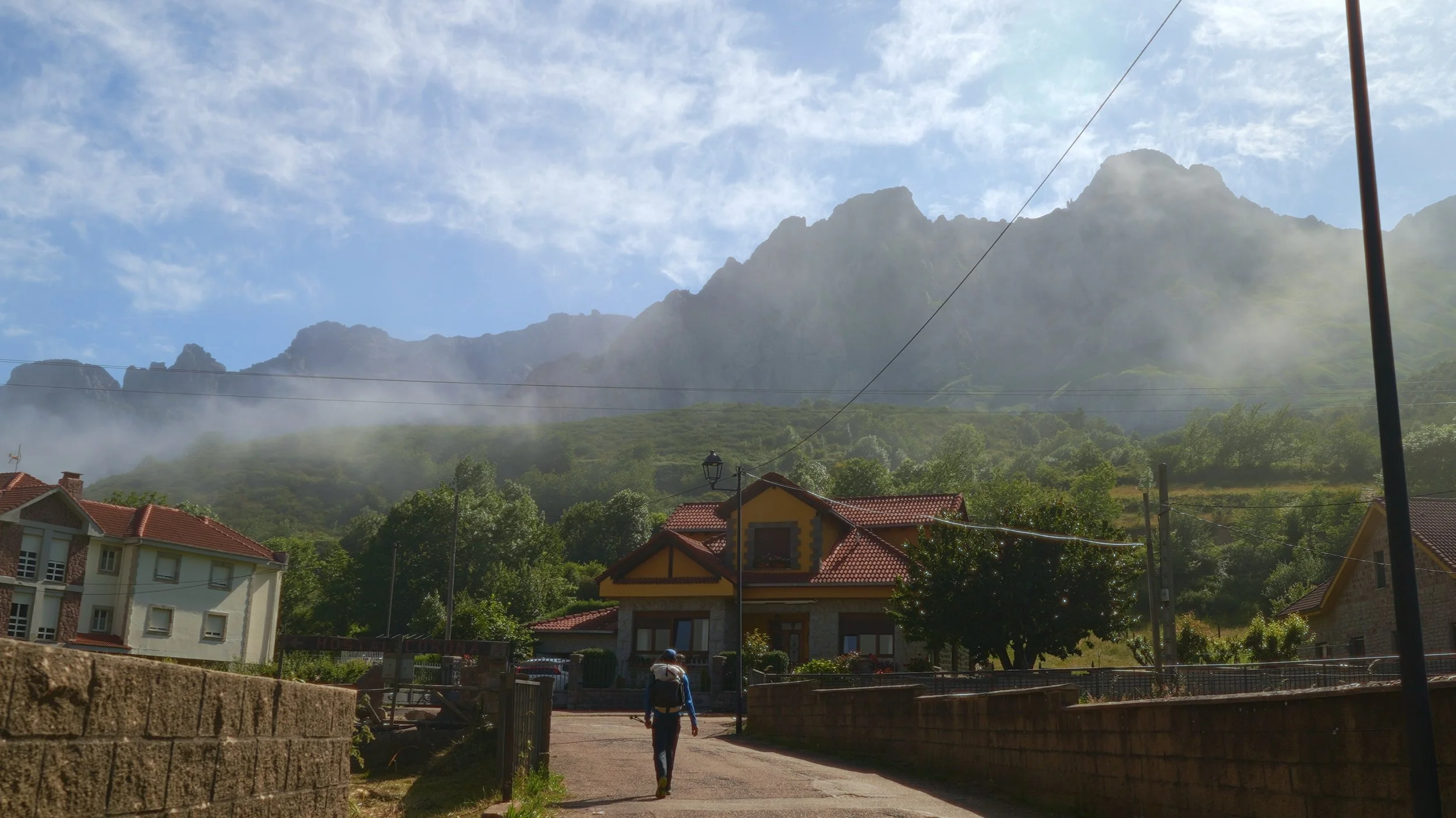 A person walking on a rural street towards a house with mountains and mist in the background.