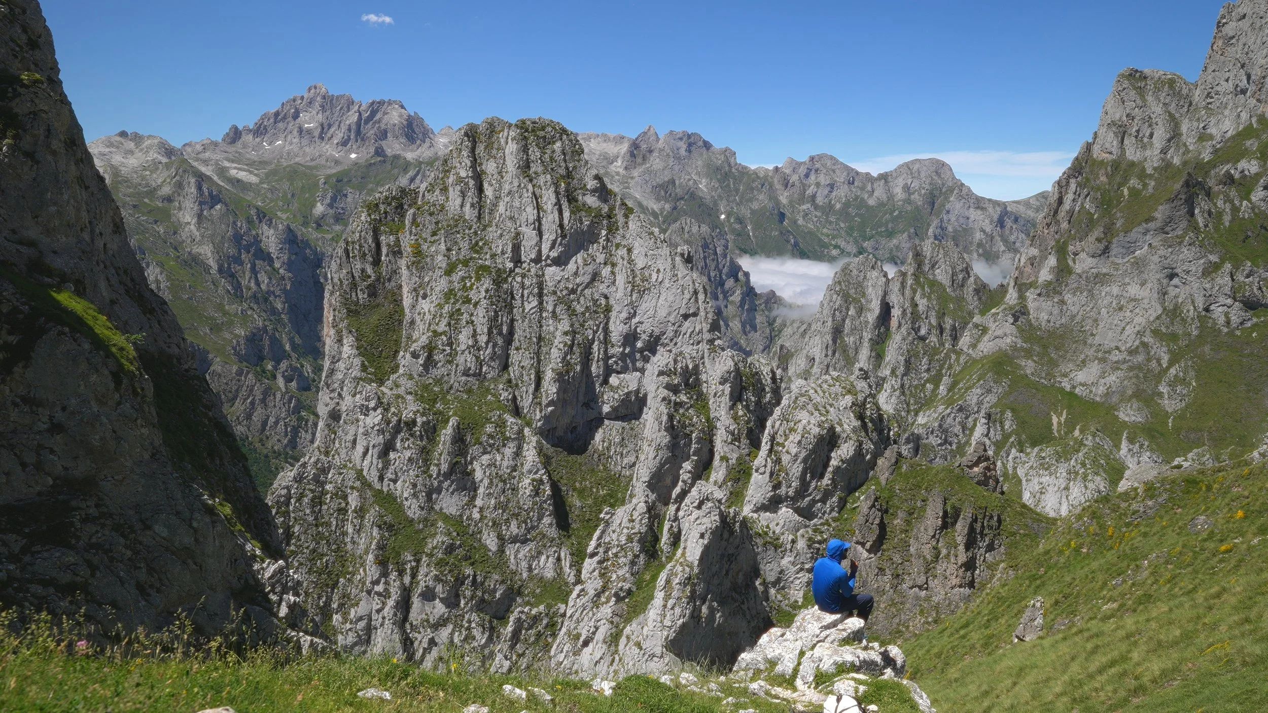A person in a blue jacket sitting on a rock in a green valley surrounded by tall rocky mountains under a clear blue sky.