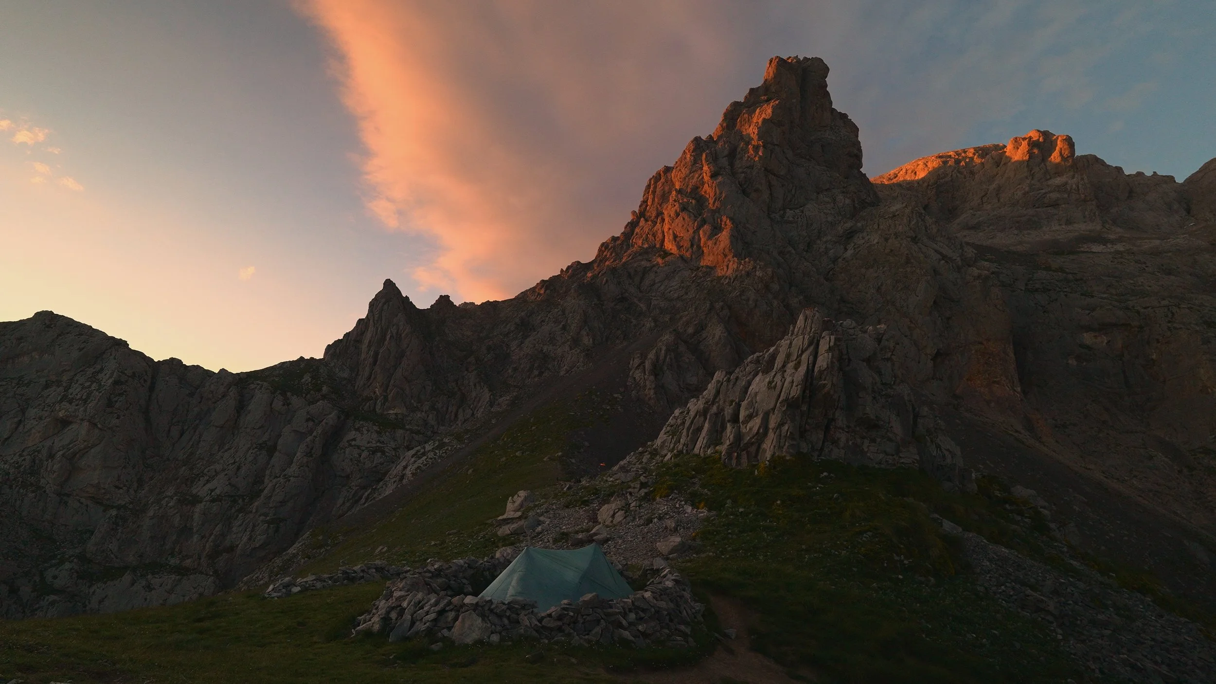 Tent set up on a grassy area at the base of a mountain during sunset, with rugged mountain peaks lit by the sunset in the background.