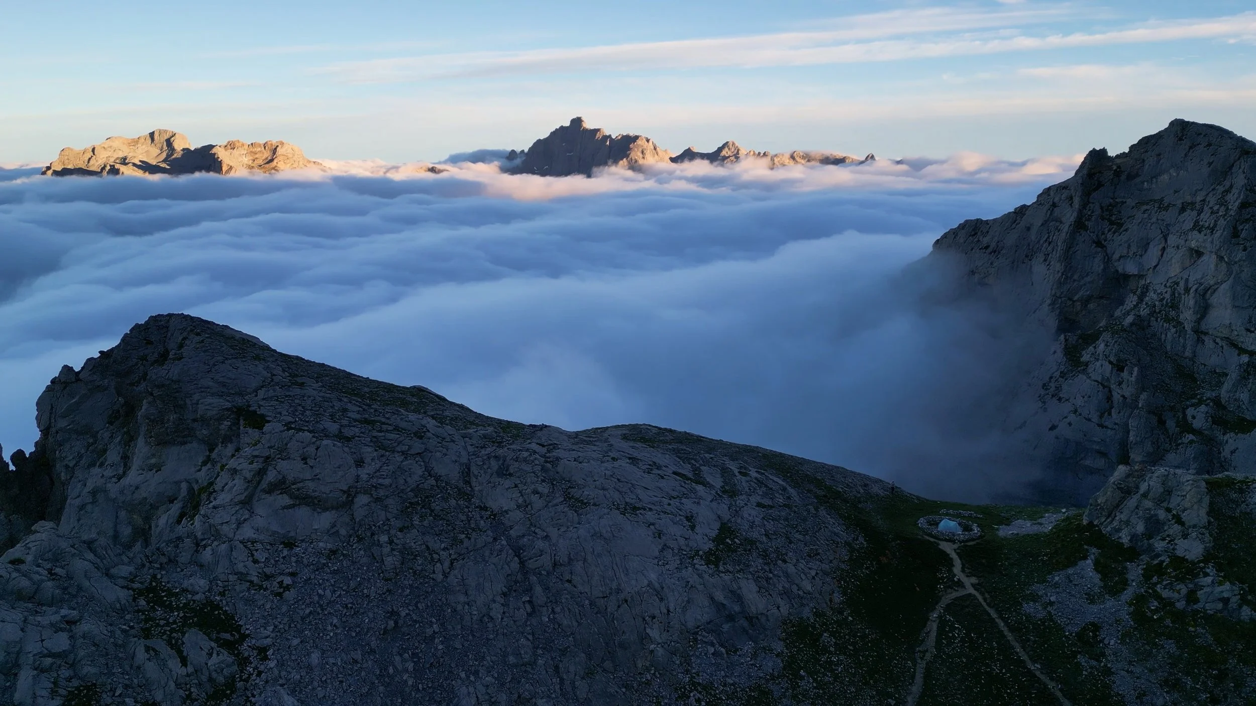 Mountain peaks above a sea of clouds during sunrise or sunset with a small tent near a rocky trail in the foreground.